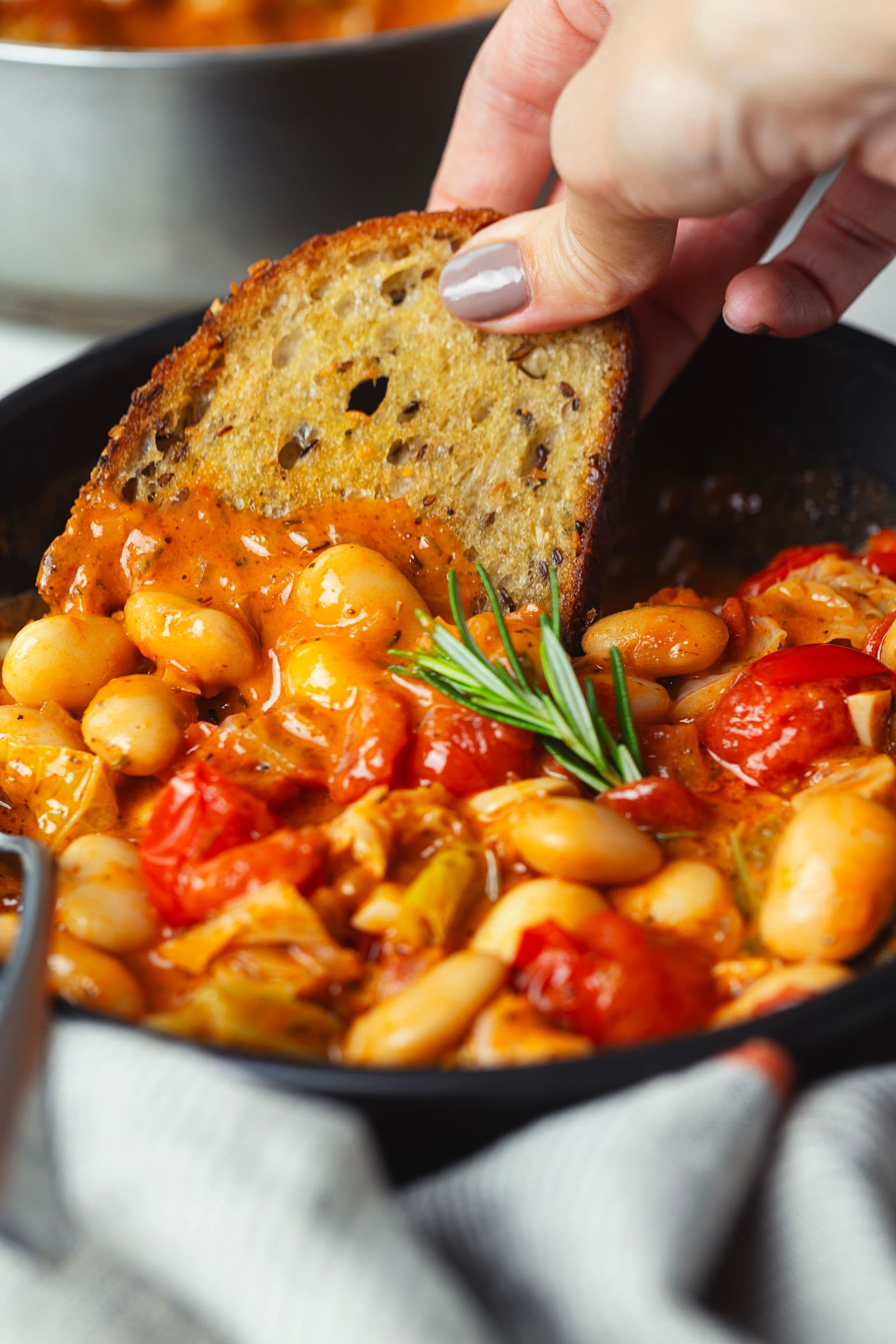 A hand dipping toasted bread into a bowl of creamy bean and tomato stew.