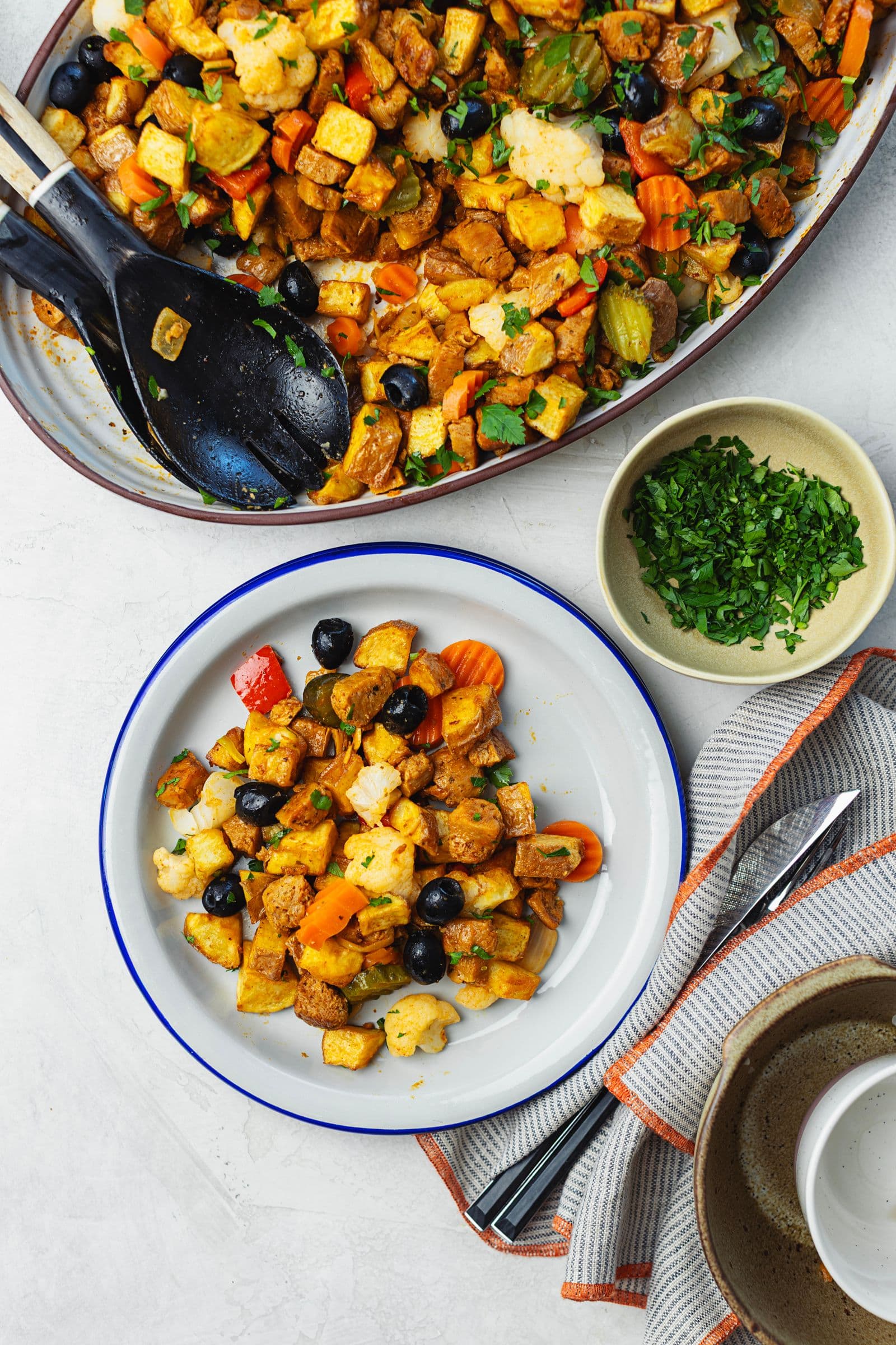 Serving platter of soy pica pau in the background with serving spoons, and a blue-rimmed individual plate with a served portion in the foreground, alongside chopped parsley