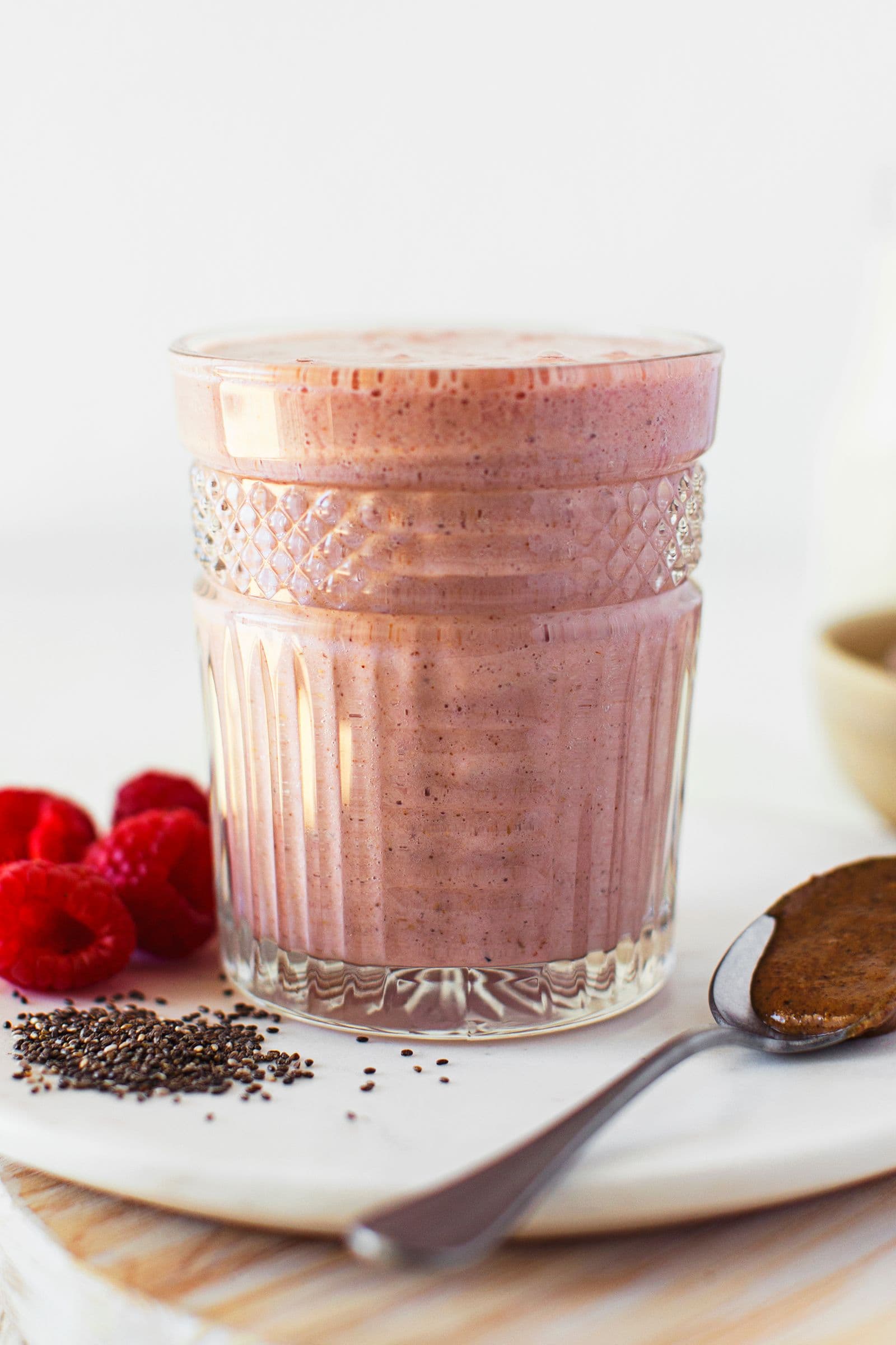 Glass of pink smoothie on a plate with chia seeds, raspberries and a spoon.