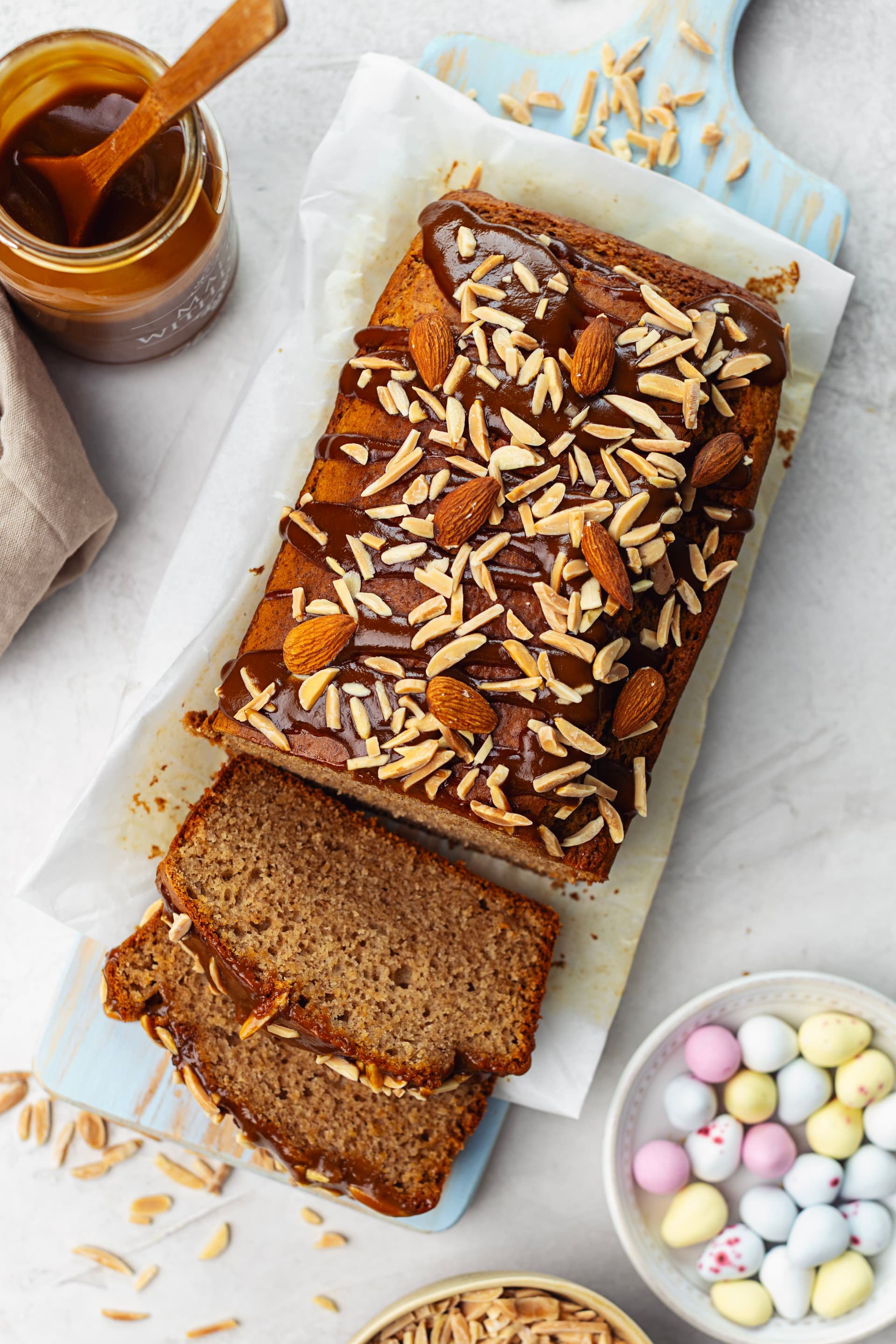 Partially sliced almond cake with coconut caramel on a light blue board, with a caramel jar and pastel Easter eggs.
