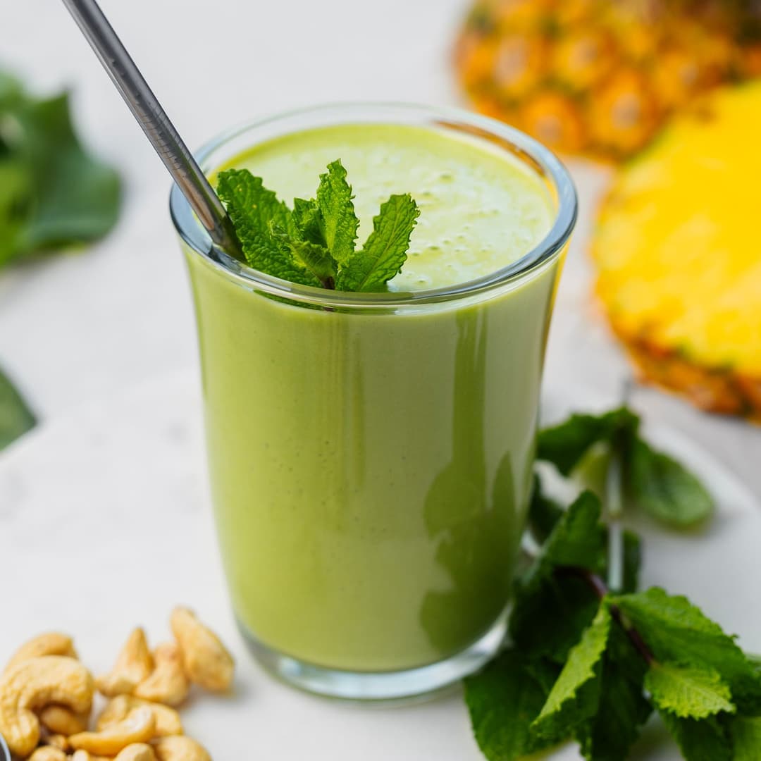 Creamy green smoothie in a glass with a metal straw and mint, surrounded by cashews, spinach, and pineapple.