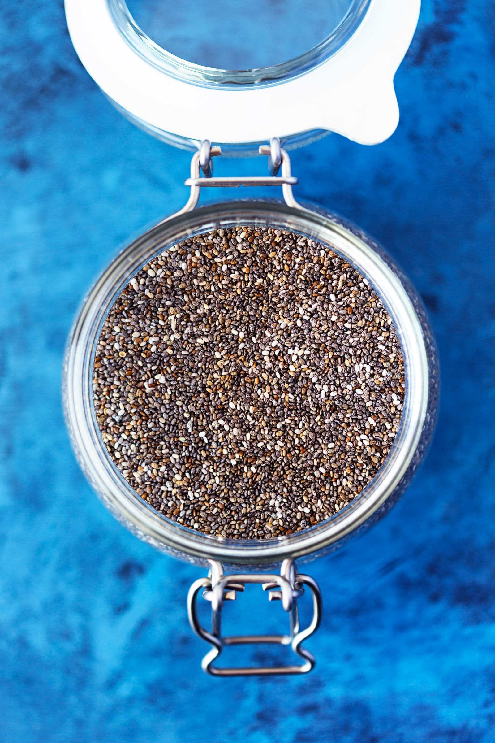 Glass jar filled with dry chia seeds on a blue surface.