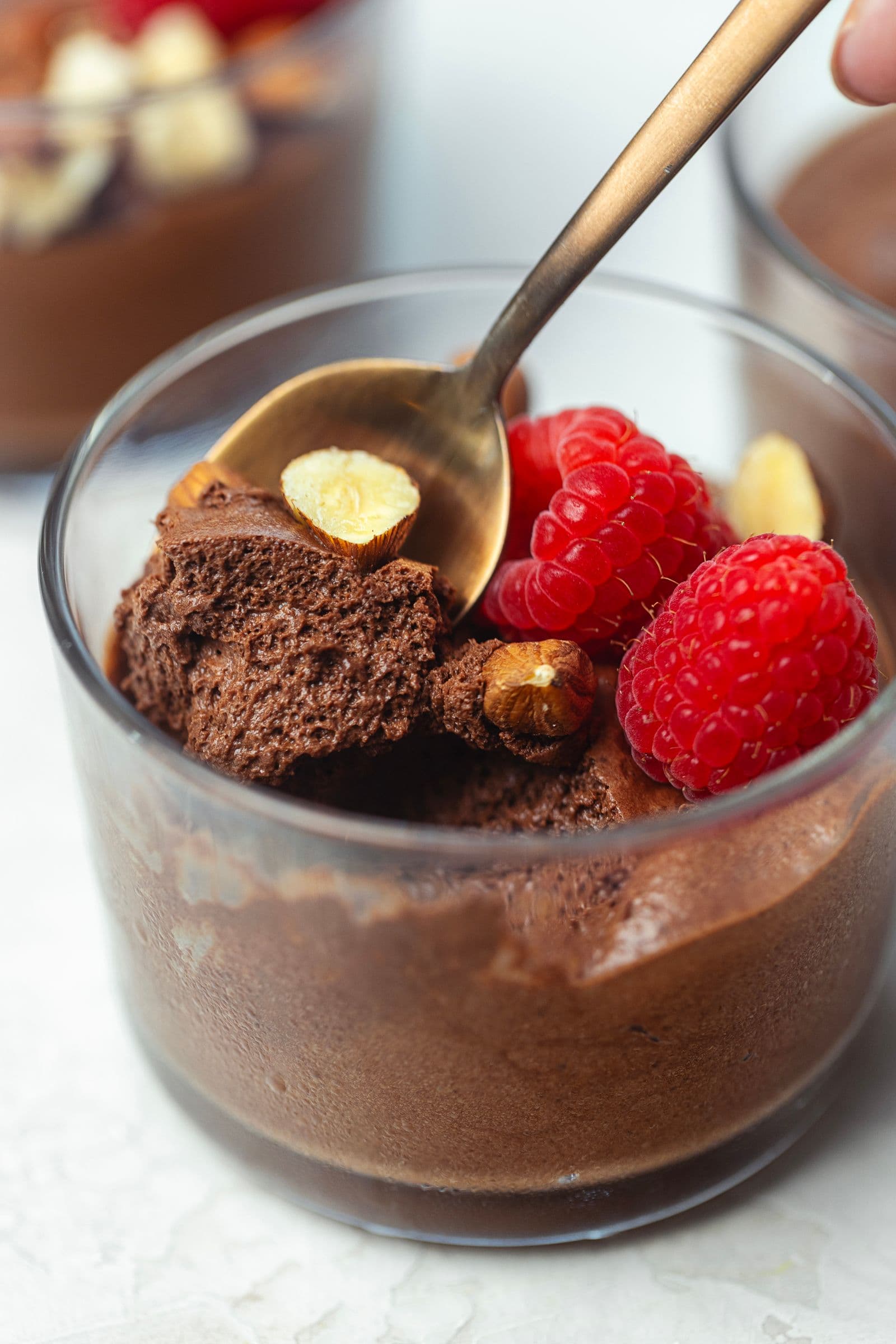 Spoon scooping a portion of fluffy, airy vegan chocolate mousse from a glass cup topped with raspberries and hazelnuts.