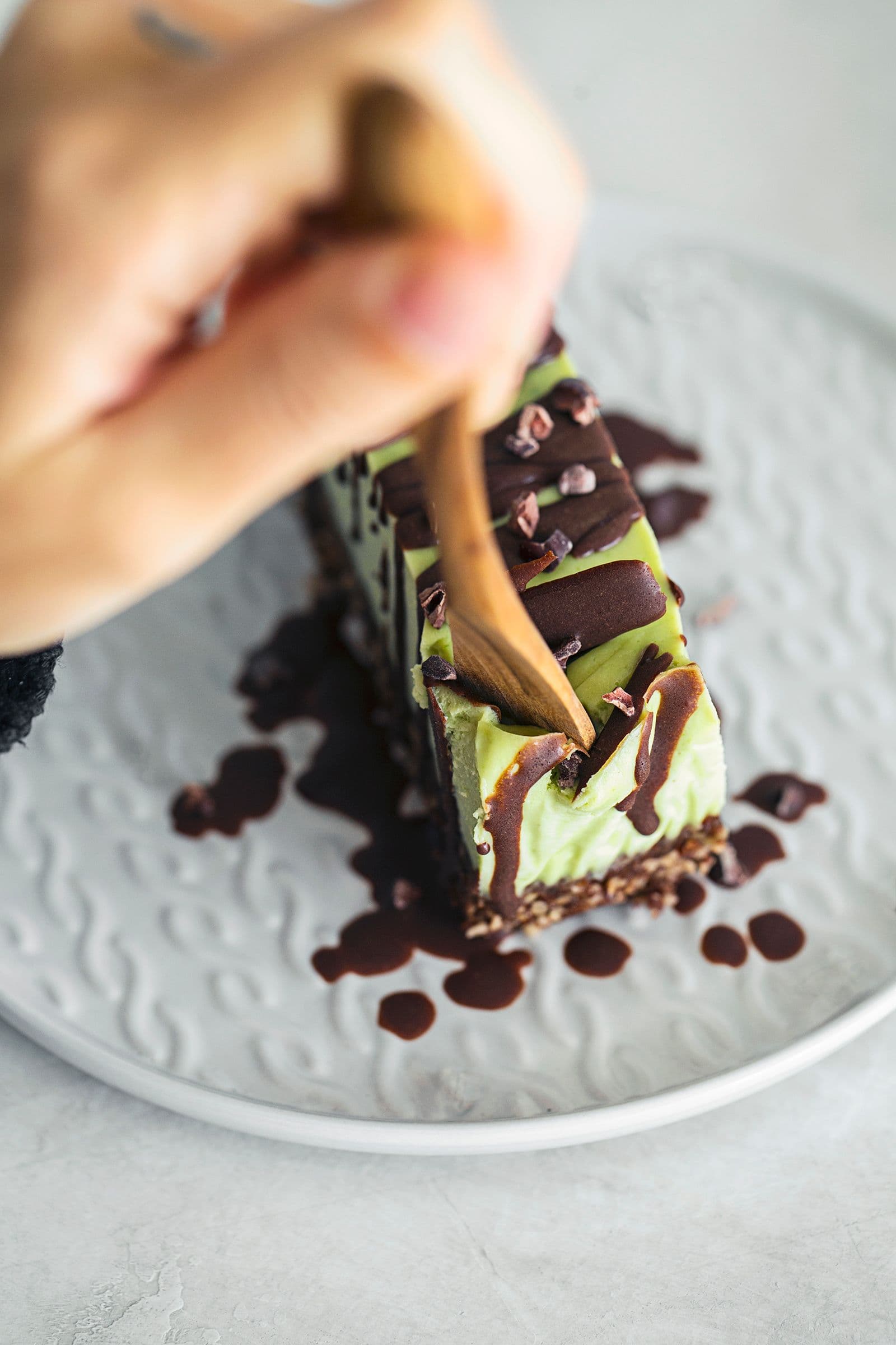Handing holding fork and digging into a slice of vegan mint and chocolate cheesecake with a nutty base, topped with melted chocolate and cacao nibs.
