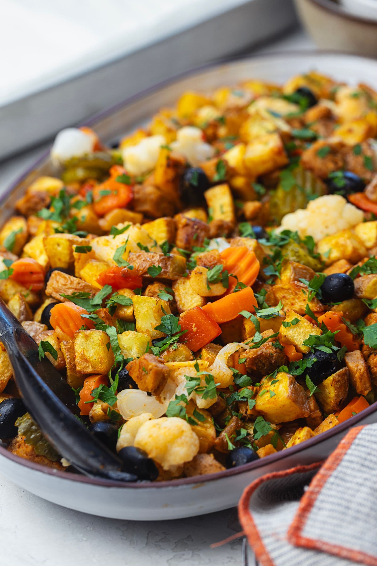 Serving dish of soy pica pau scattered with fresh chopped parsley, featuring soy, potatoes, pickles, olives and carrots, shot at a slight angle