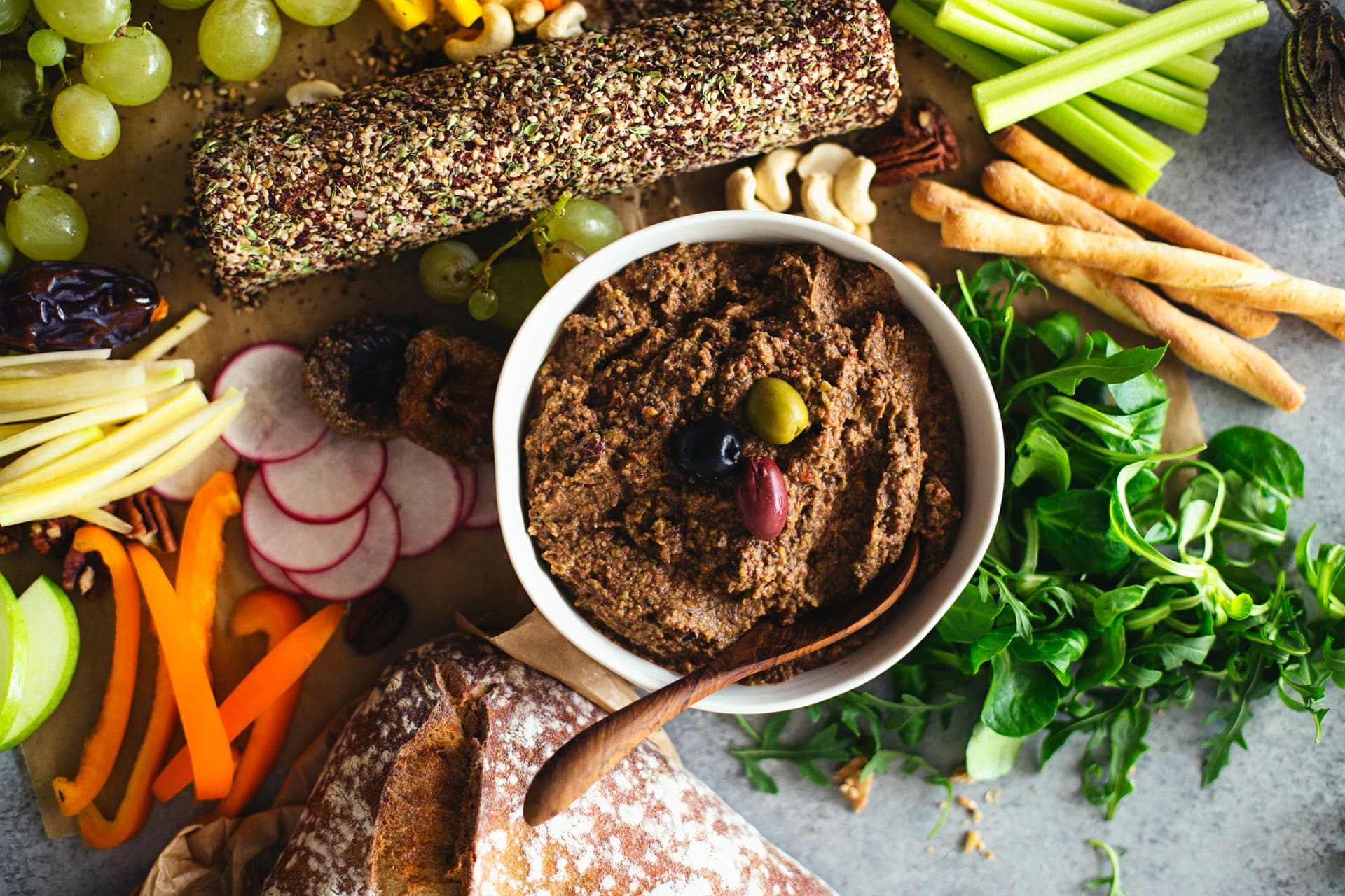 Cashew cheese log and lentil pâté served on a large platter with grapes, nuts, raw vegetables and bread slices.