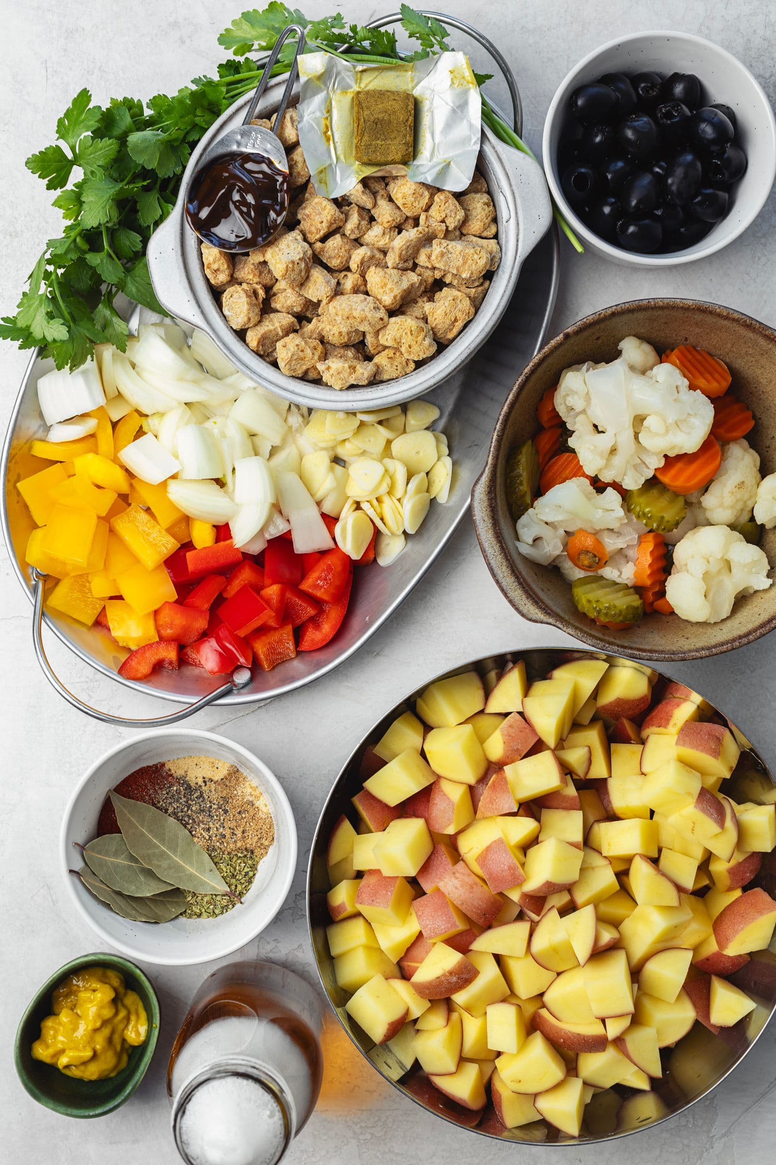 Flat lay of soy pica pau ingredients in bowls: dry soy chunks, cubed red potatoes, bell peppers, onion, garlic, black olives, pickled vegetables, spices, mustard and fresh parsley