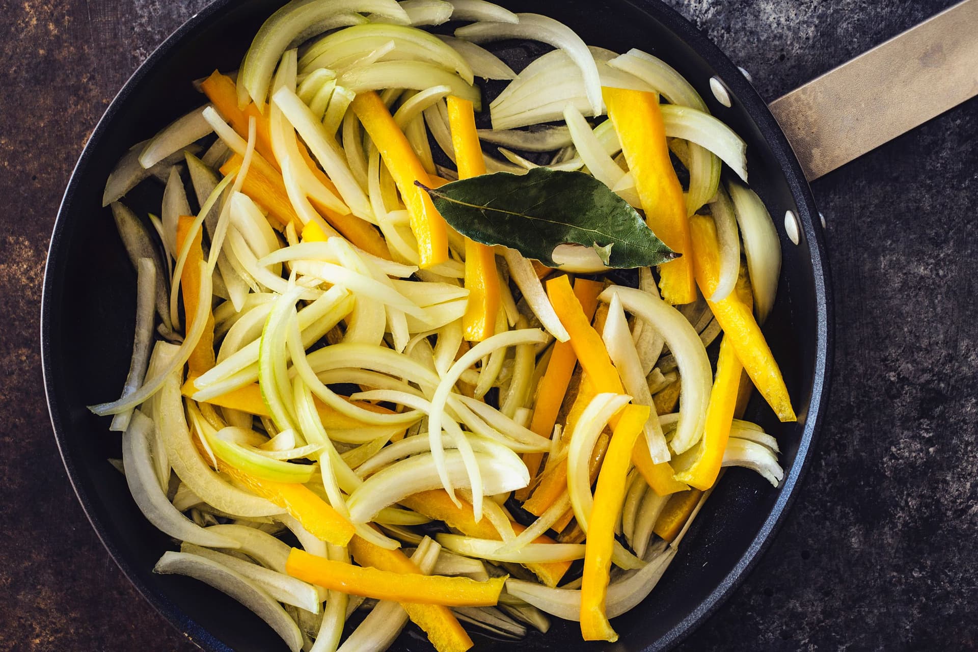 A black skillet with sliced onions, yellow bell peppers, and a bay leaf sautéing.