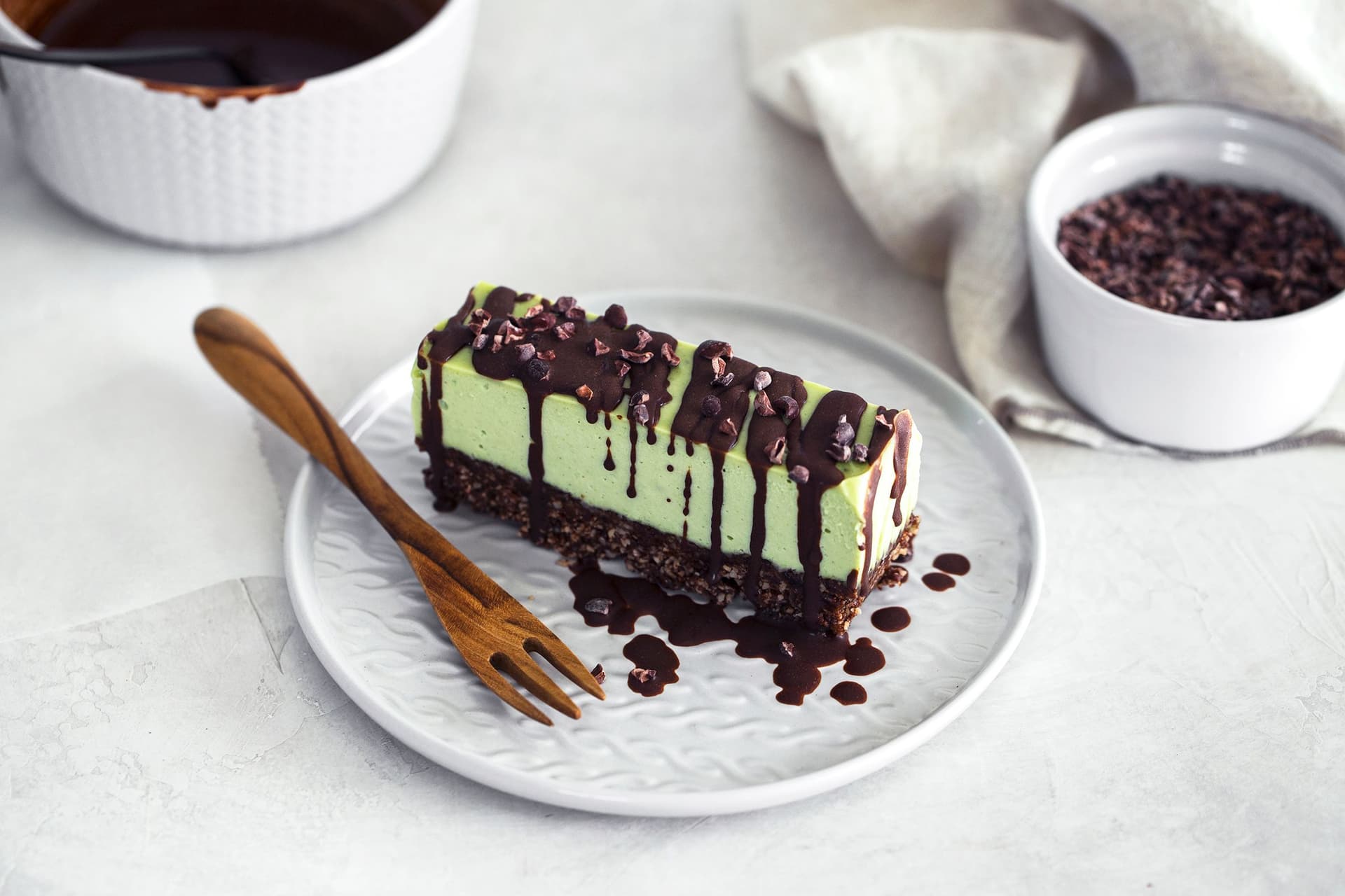Slice of vegan mint and chocolate cheesecake with a nutty base, topped with melted chocolate and cacao nibs.