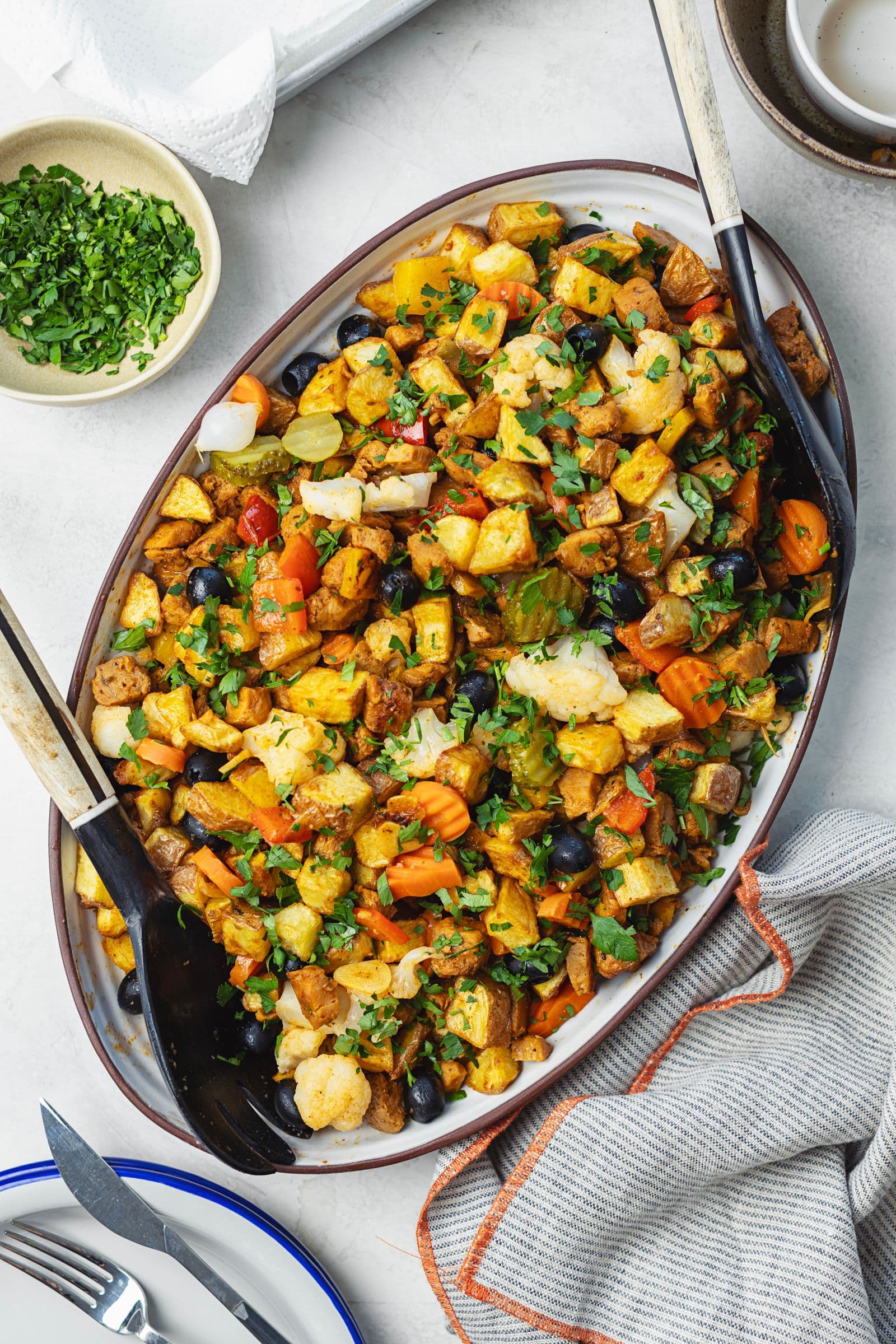 Overhead shot of oval platter with complete soy pica pau topped with chopped parsley, parsley bowl and cutlery alongside