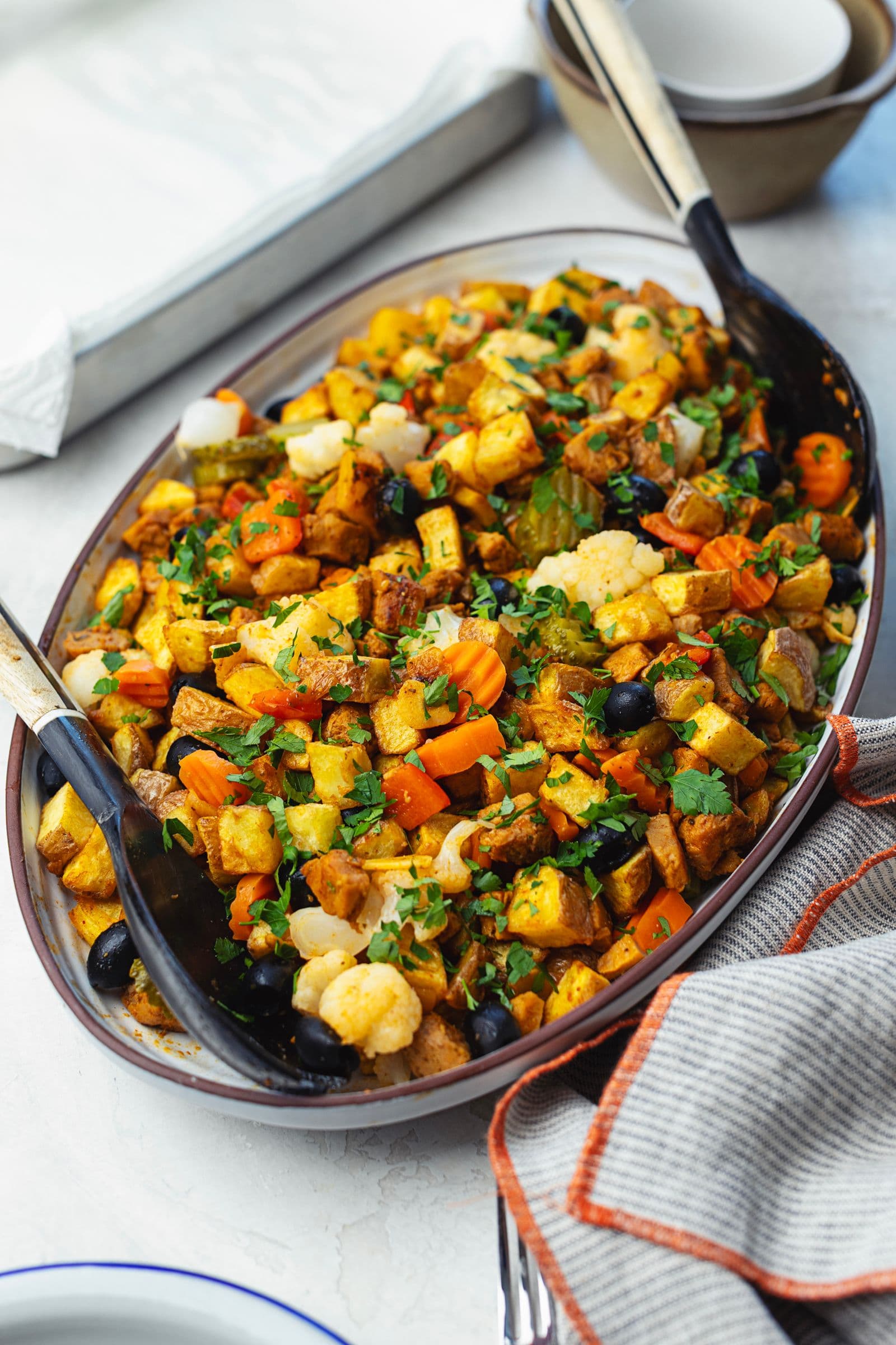 Oval platter of soy pica pau with crispy potatoes, black olives, pickles, carrot and cauliflower, scattered with fresh parsley, shot from a slight side angle