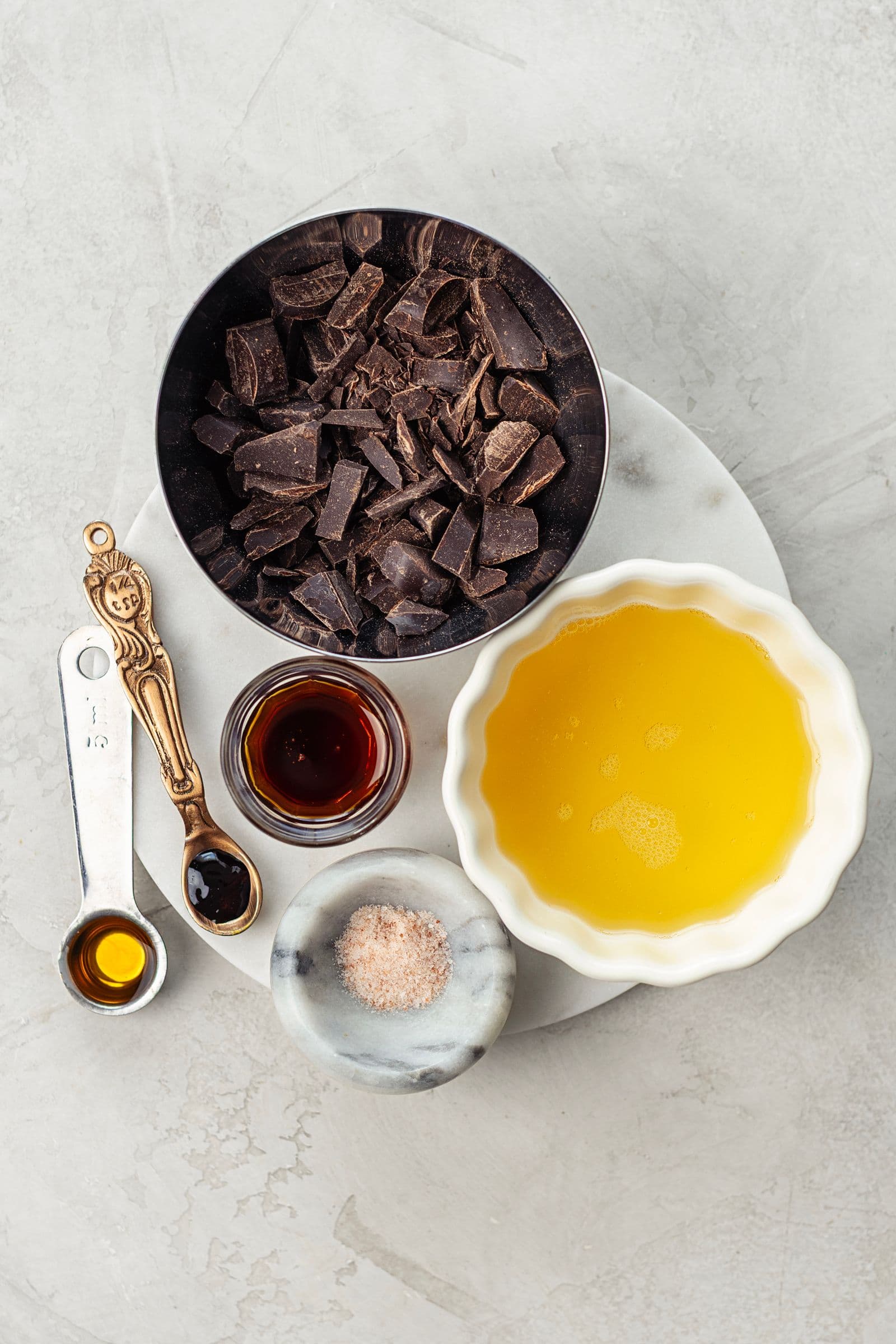 Ingredients for vegan chocolate mousse arranged on a light surface, including chopped dark chocolate, aquafaba, maple syrup, vanilla and salt.
