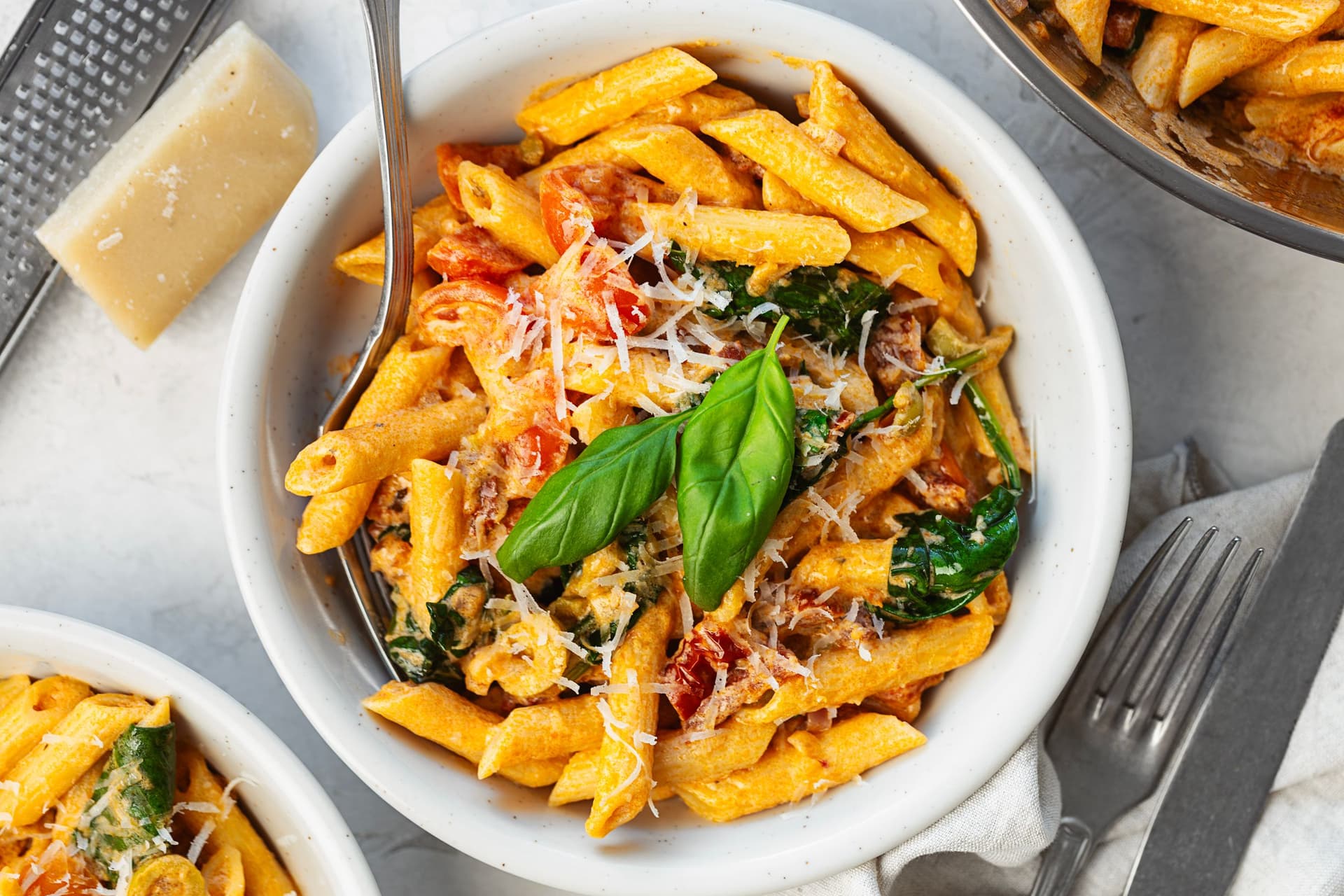 Top-down view of a bowl of creamy tomato and spinach penne next to the serving pan, garnished with fresh basil