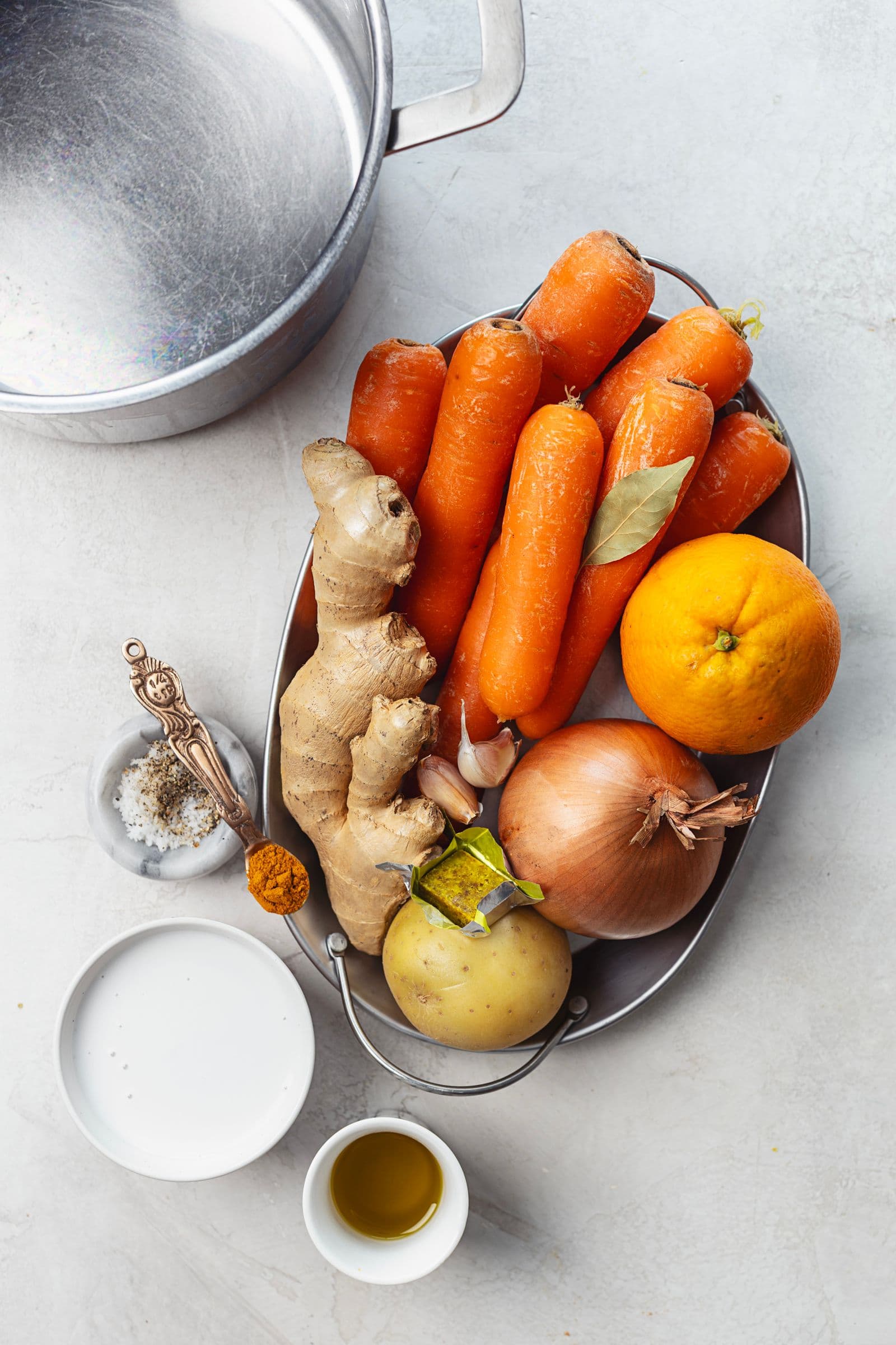 Ingredients for creamy carrot soup arranged on a tray: carrots, ginger, onion, garlic, orange, potato, olive oil, spices, and coconut milk beside an empty pot.