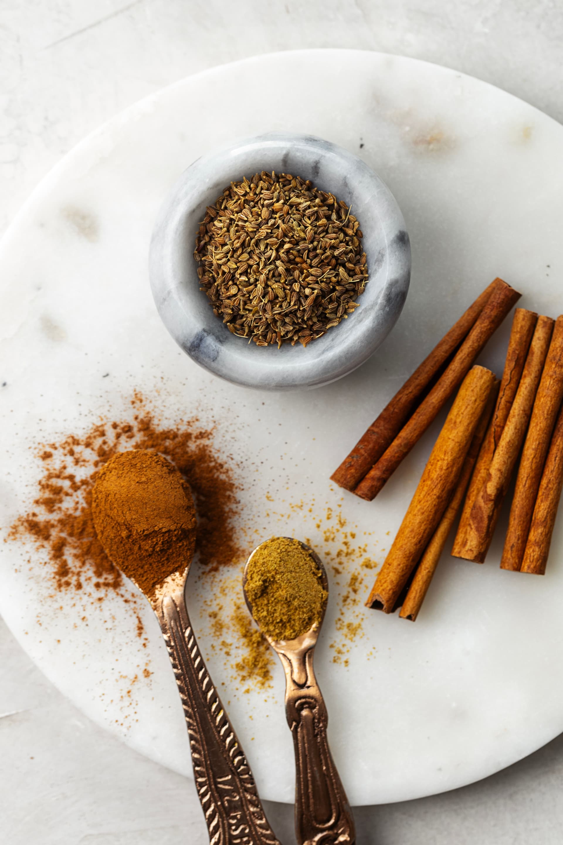 Folar spices on a marble board: whole anise seeds, cinnamon sticks, and spoons of ground cinnamon and anise.
