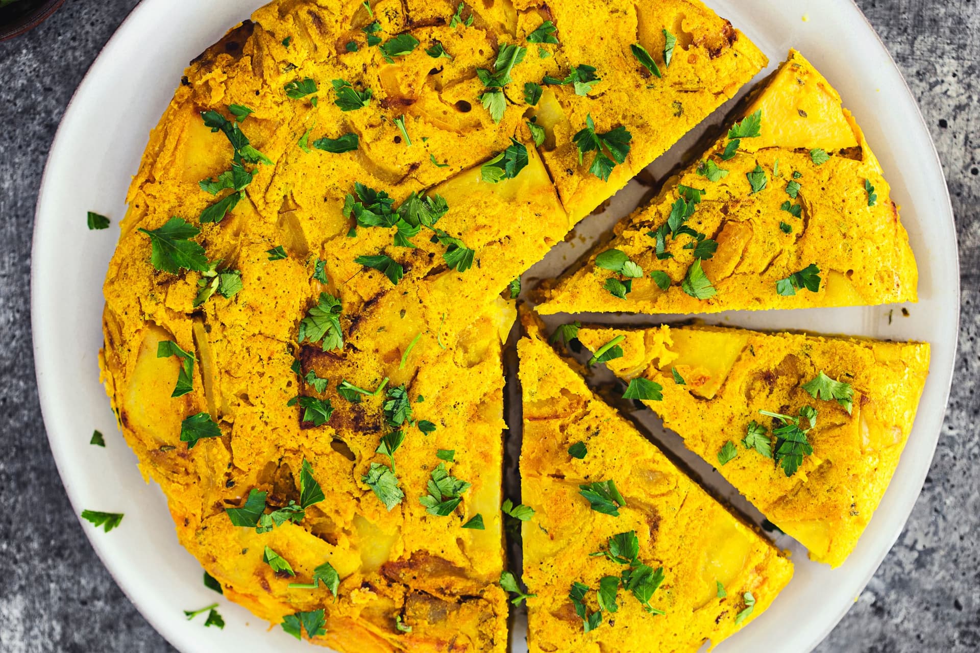 Vegan Spanish tortilla on a white plate, garnished with parsley, with three slices cut.