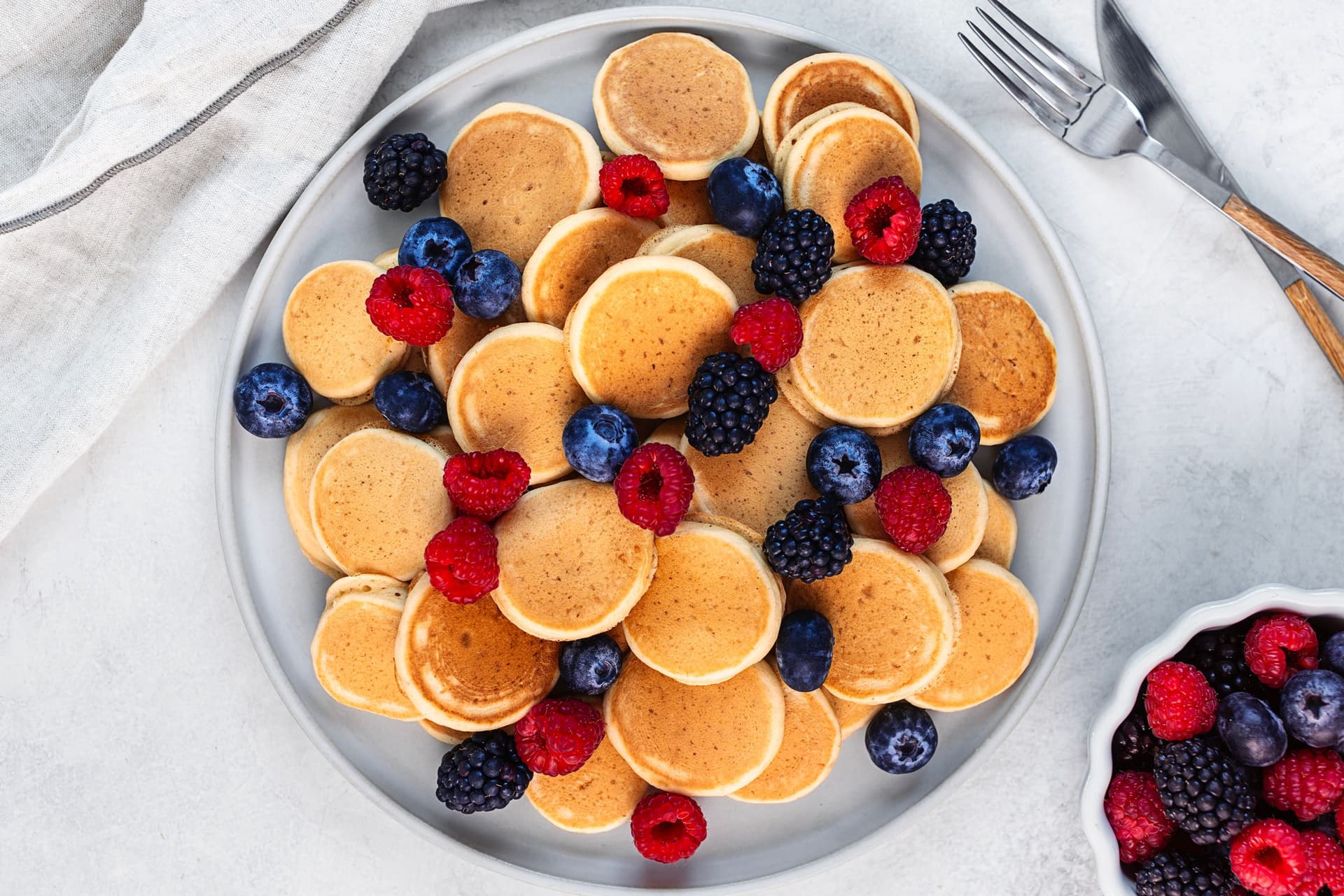 Plate of mini vegan pancakes topped with fresh raspberries, blueberries, and blackberries, with cutlery on the side