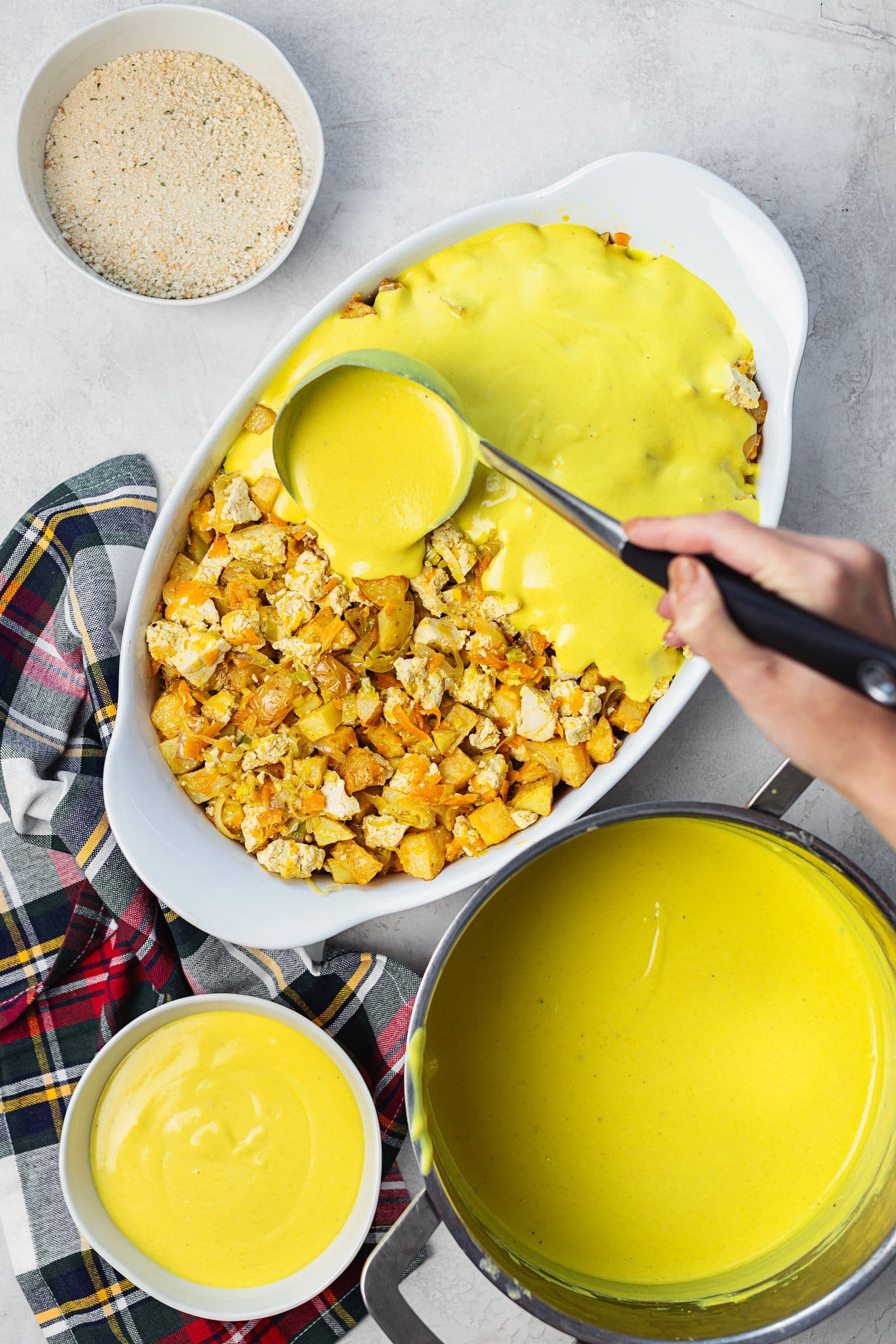 Yellow béchamel sauce being ladled over the tofu, vegetable and potato mixture in the baking dish.