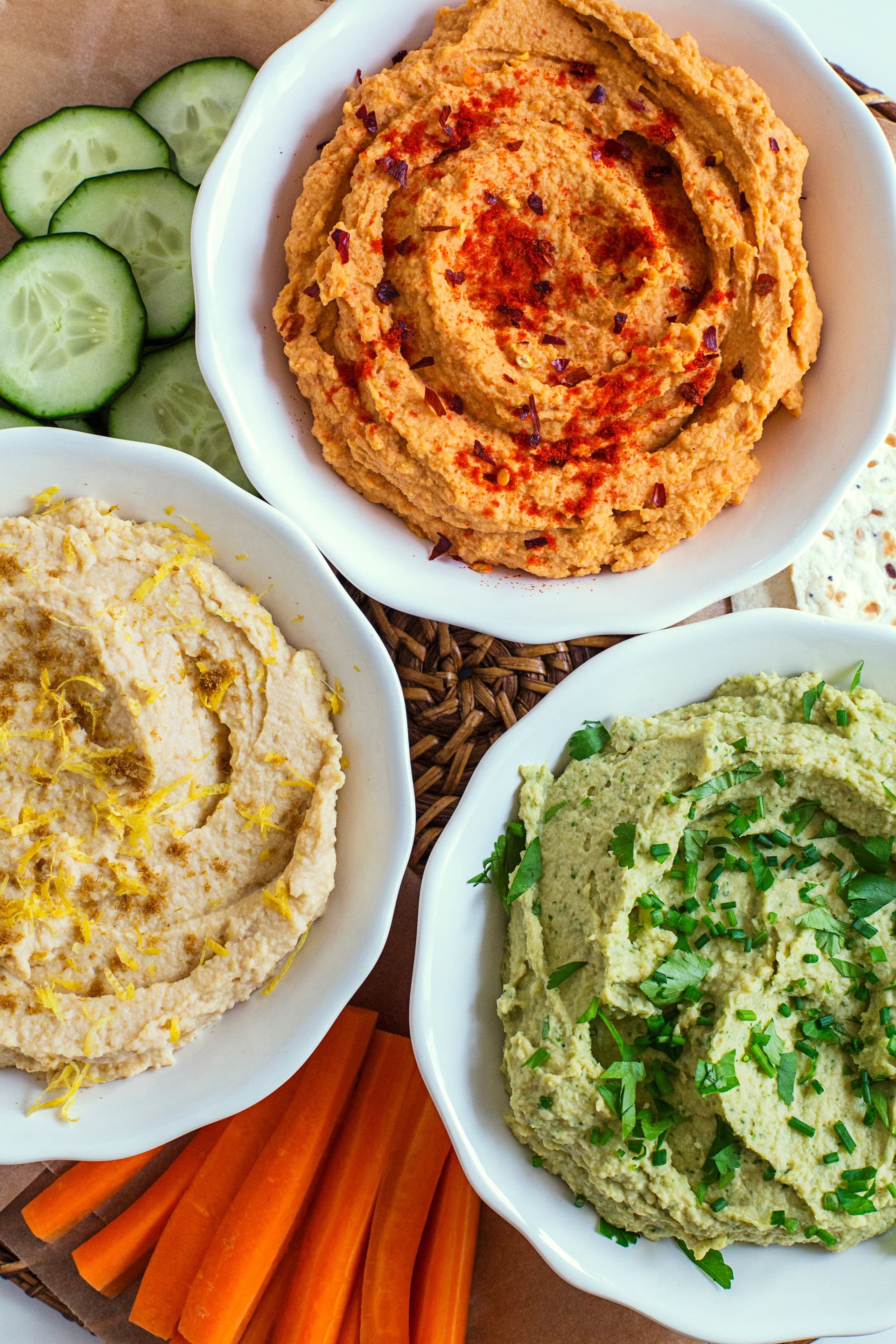 Three colourful bowls of hummus—lemon, tomato, and herb—served with fresh tortilla and vegetable sticks.
