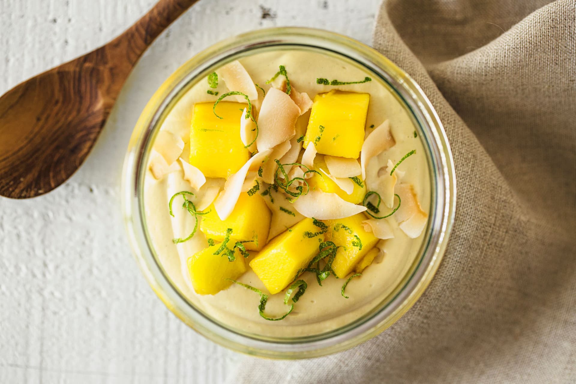 Glass bowl of vegan mango cashew yogurt topped with fresh mango cubes, lime zest and toasted coconut flakes.