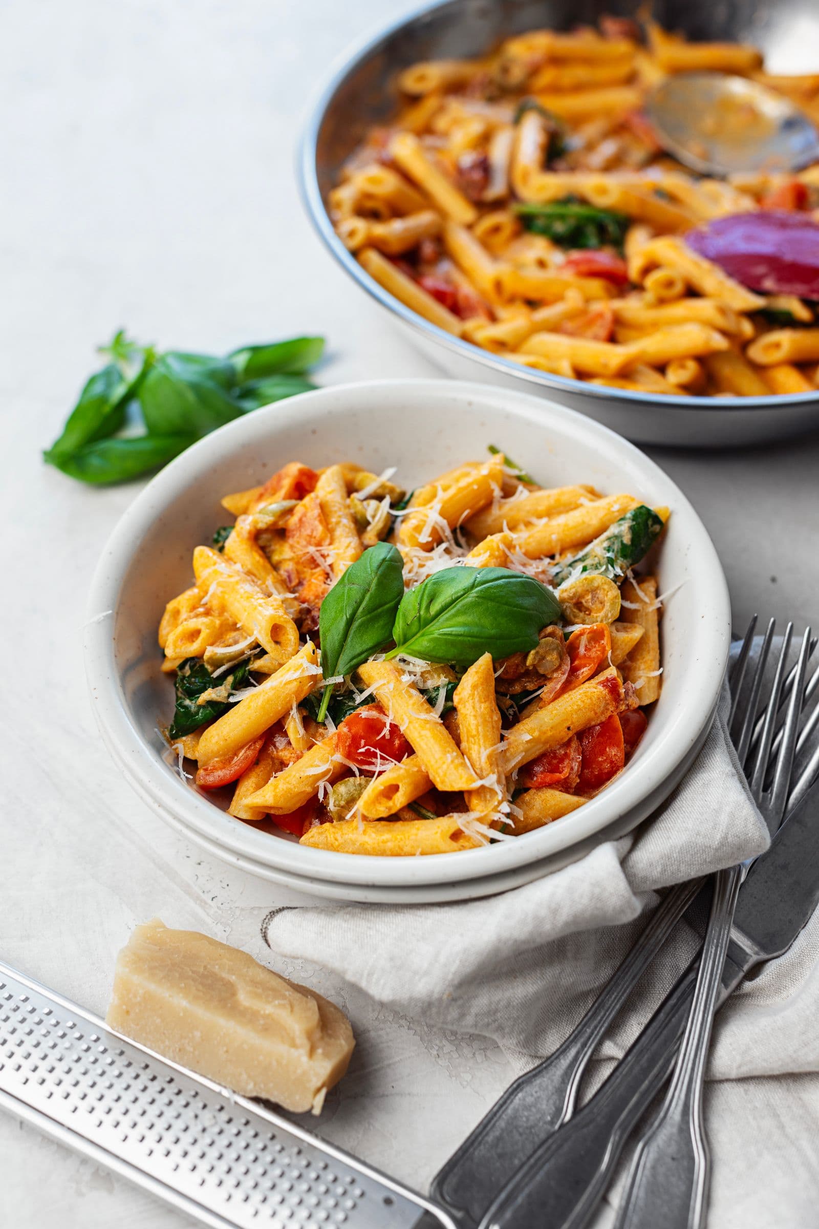 Bowl of creamy tomato and spinach penne with fresh basil and grated vegan parmesan, with the pan in the background