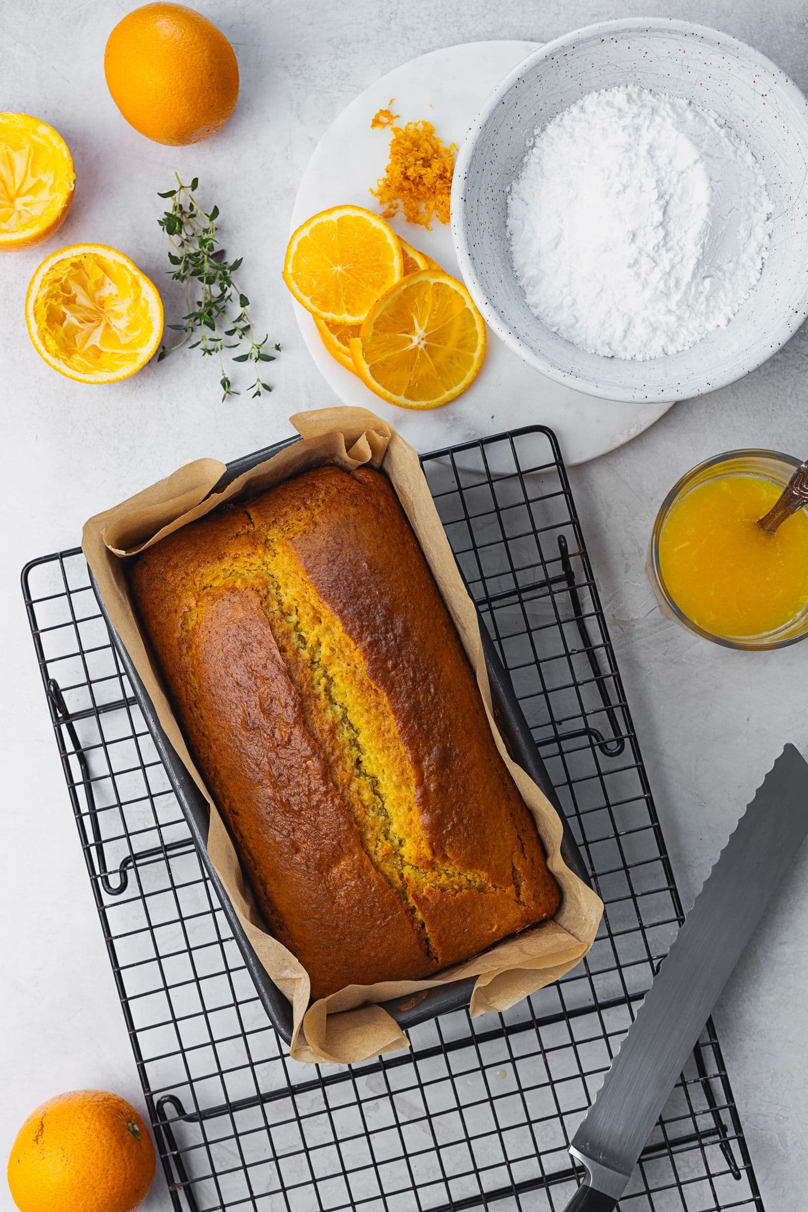 Freshly baked orange cake still in the pan on a cooling rack, with glaze ingredients on the side.