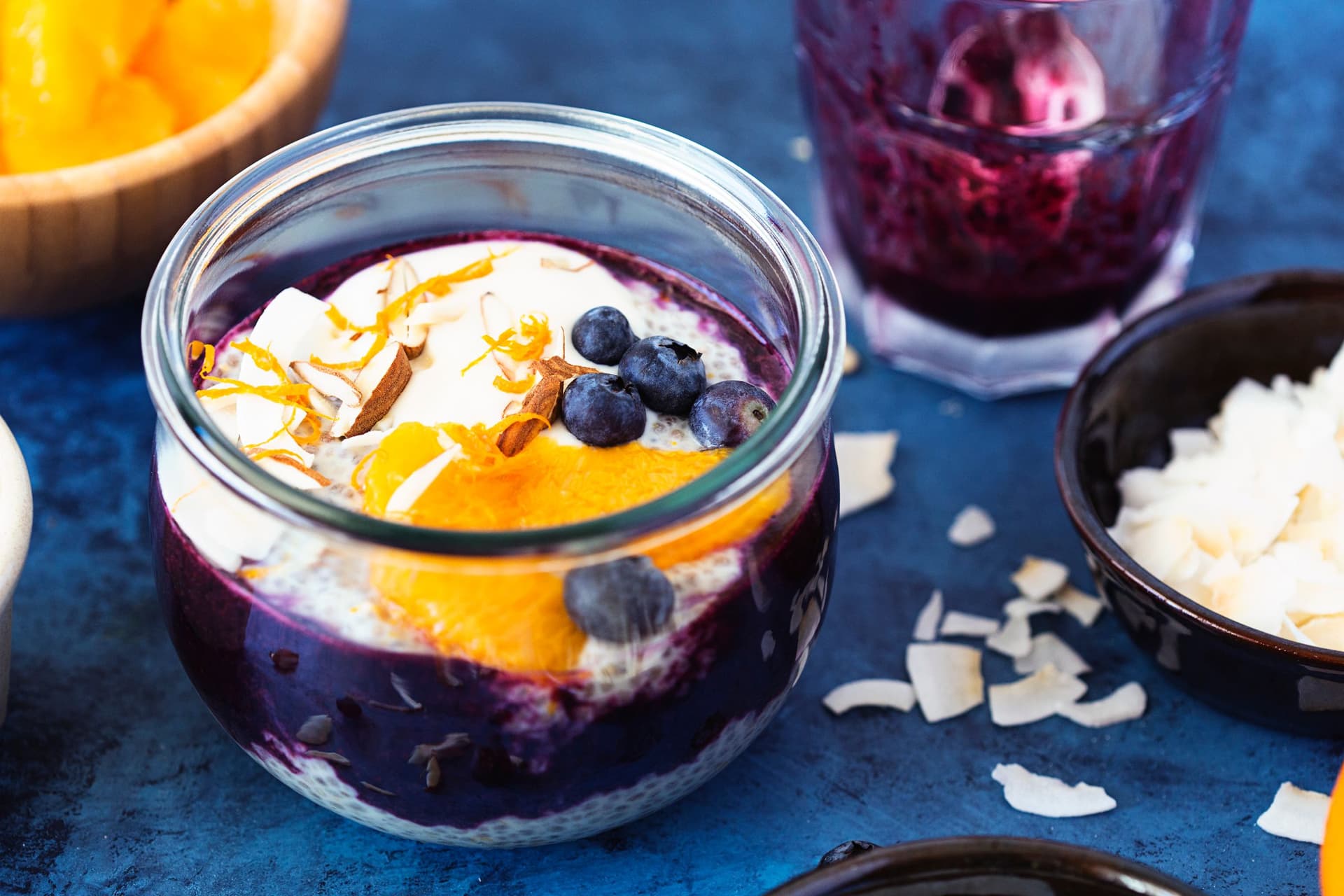 Glass jar of chia pudding layered with blueberry purée, fresh orange slices and coconut flakes.