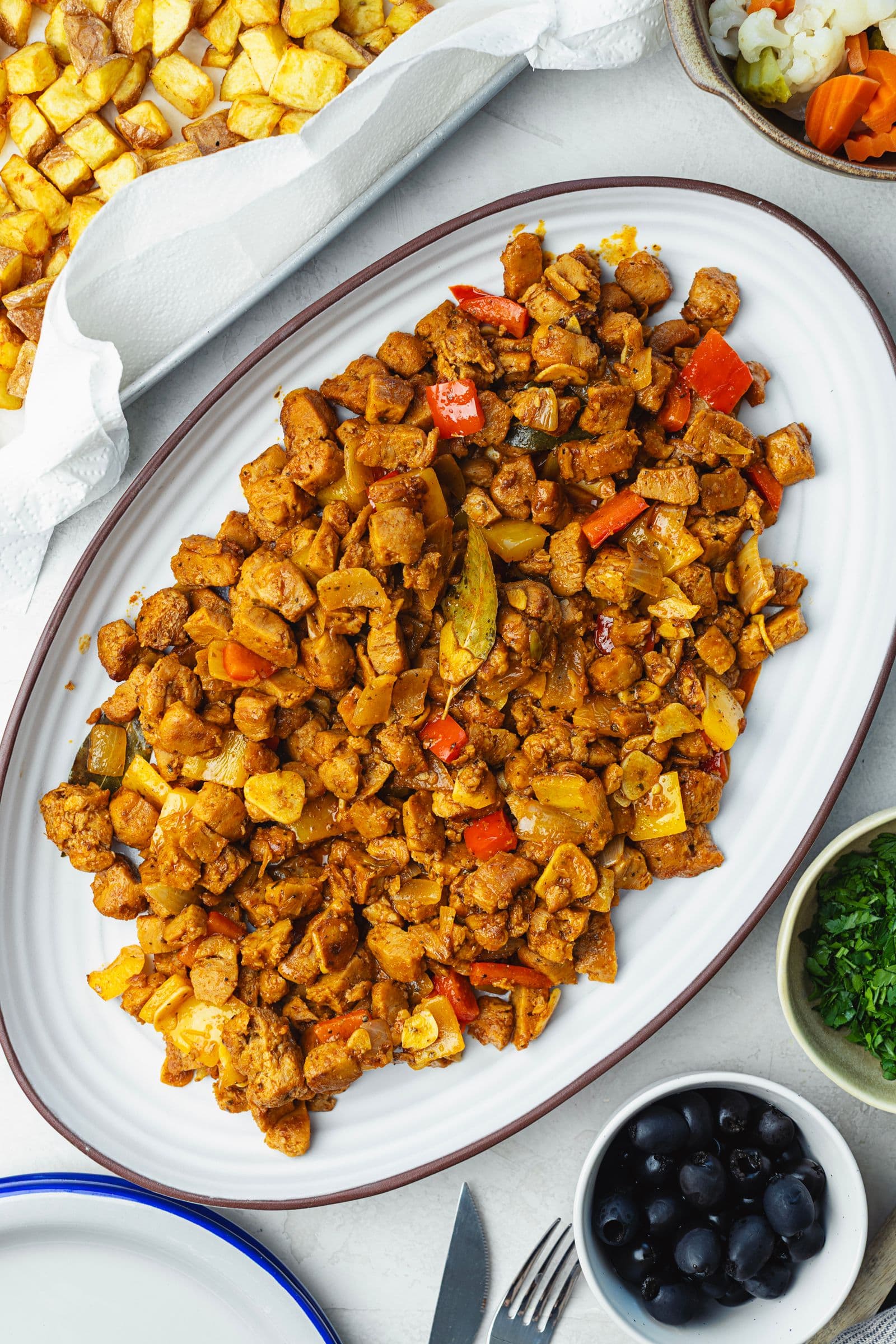 Oval serving dish with soy pica pau before adding the potatoes, surrounded by crispy potatoes, black olives and chopped parsley