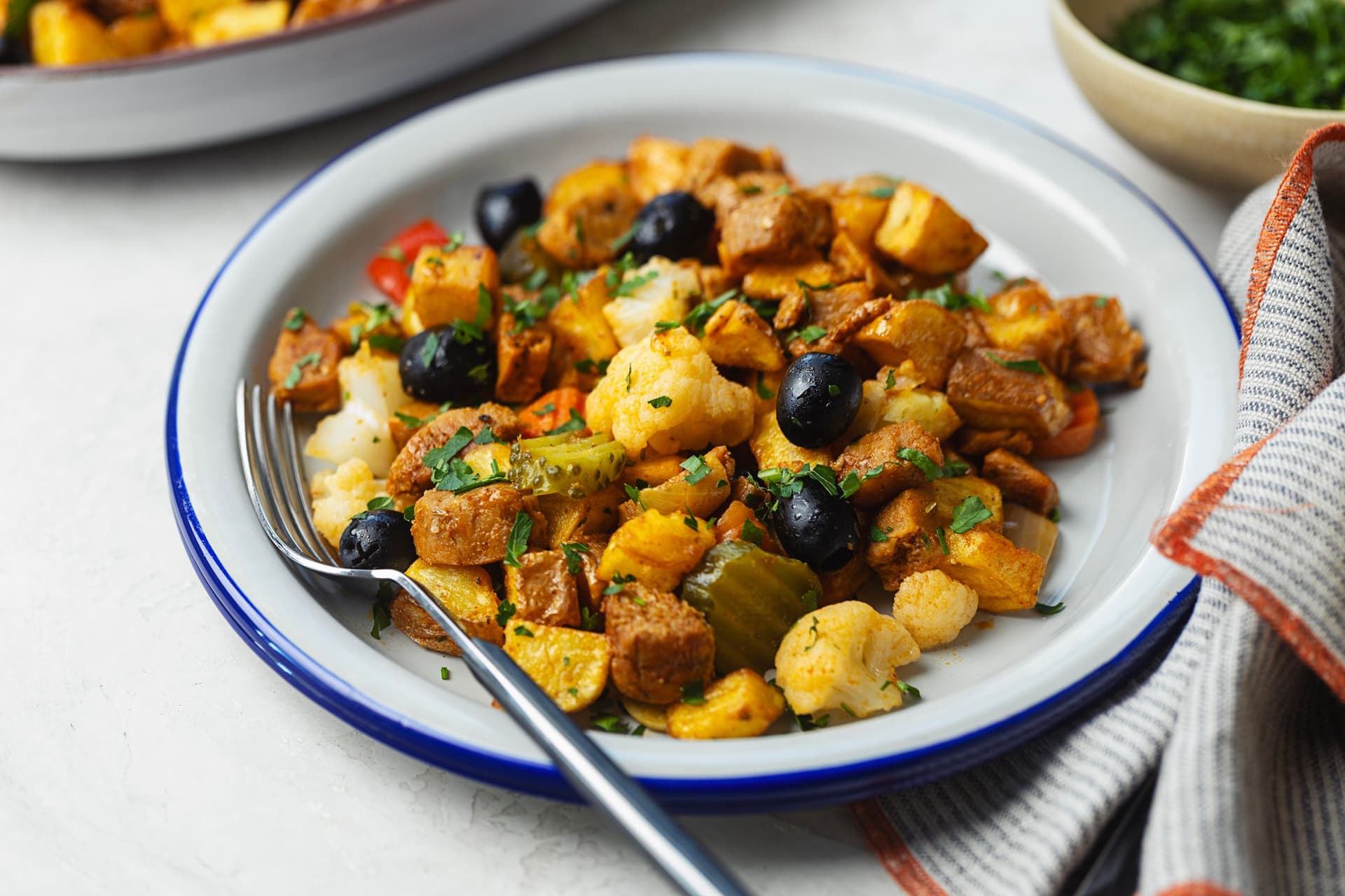 Blue-rimmed plate with a serving of soy pica pau shot from a low side angle, featuring black olives, cauliflower, pickles, crispy potatoes and caramelised soy with fresh parsley