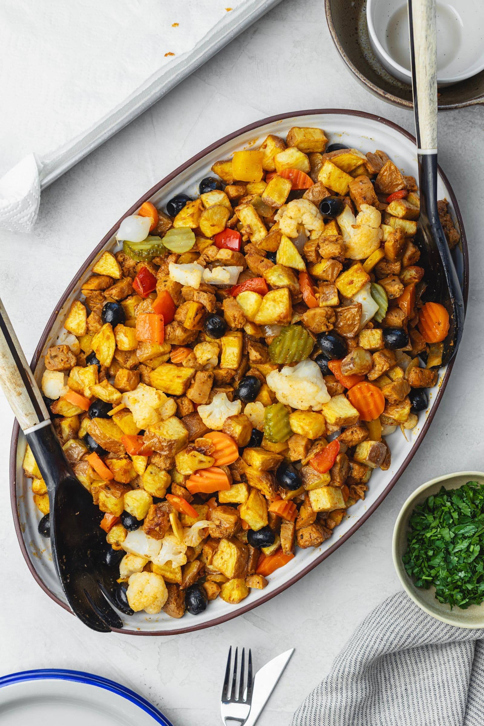 Overhead view of oval platter with complete soy pica pau: soy chunks, fried potatoes, pickles, black olives, cauliflower and carrots, with serving spoons
