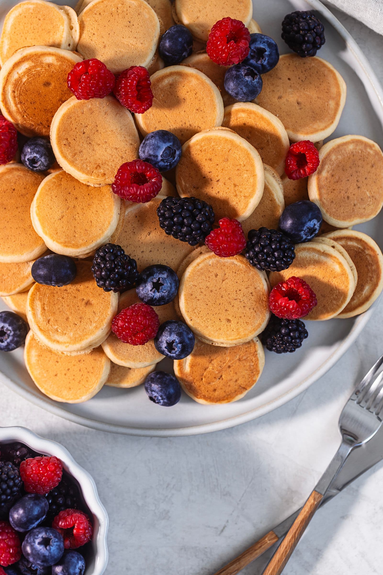 Top view of a plate of golden mini vegan pancakes topped with fresh berries, with a bowl of berries and cutlery on the side