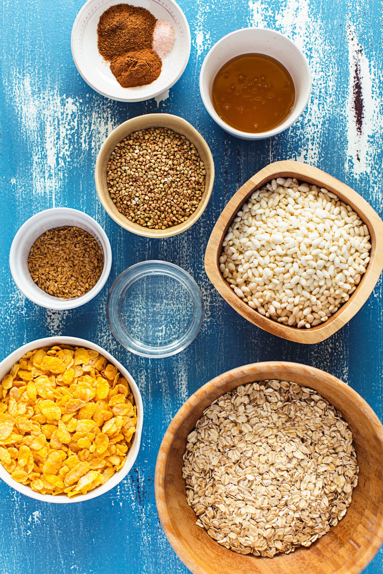 Overhead view of granola ingredients in bowls including oats, cornflakes, seeds and spices.