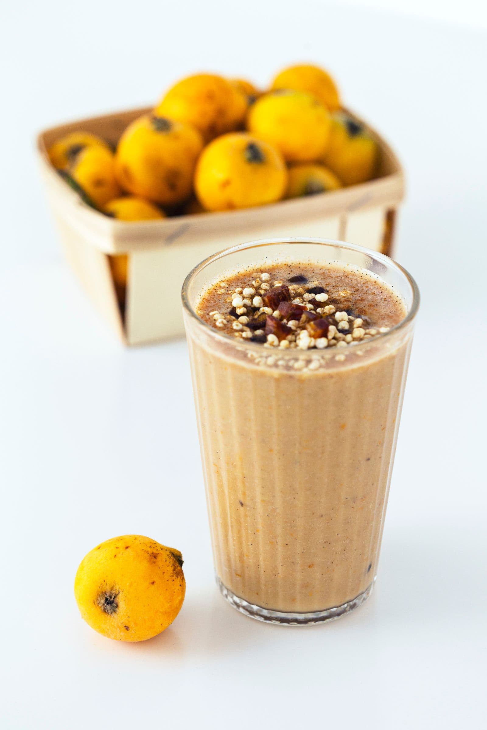 Smoothie de nêspera com topping de millet tufado e tâmara medjoul, em copo alto, ao lado de uma nêspera e cesto de fruta ao fundo