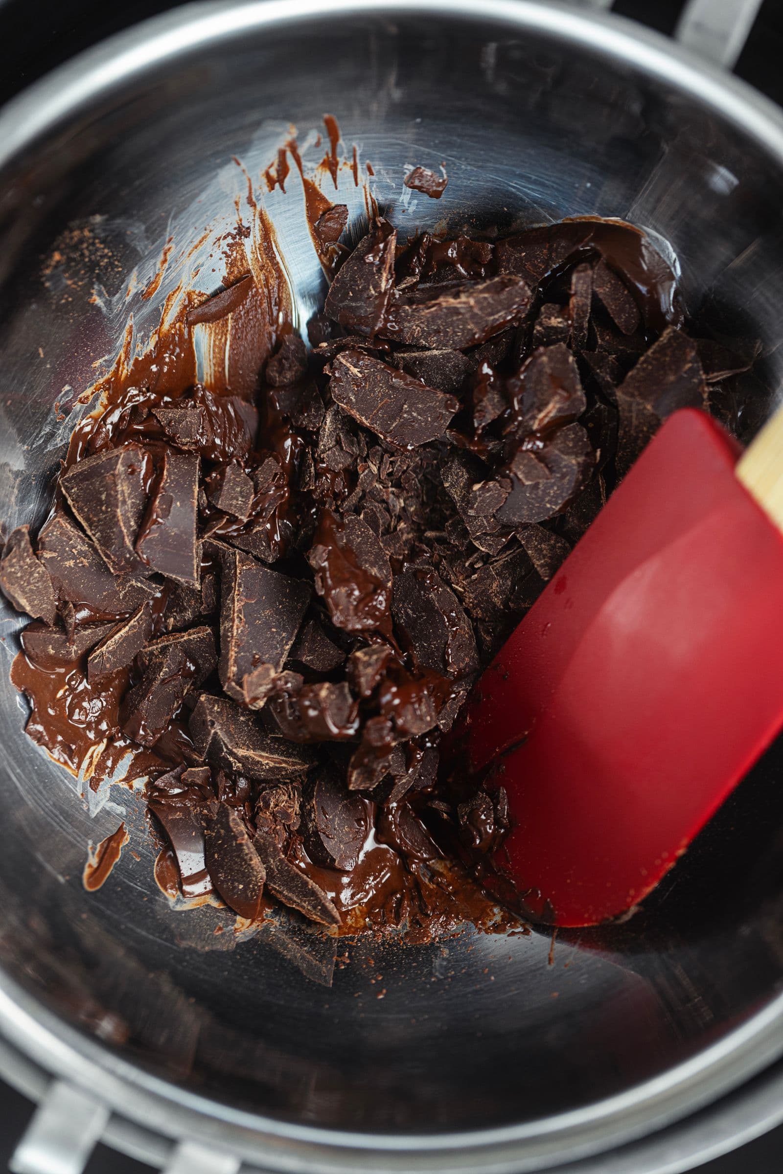 Chopped dark chocolate melting in a stainless steel bowl over a bain-marie, with a silicone spatula.