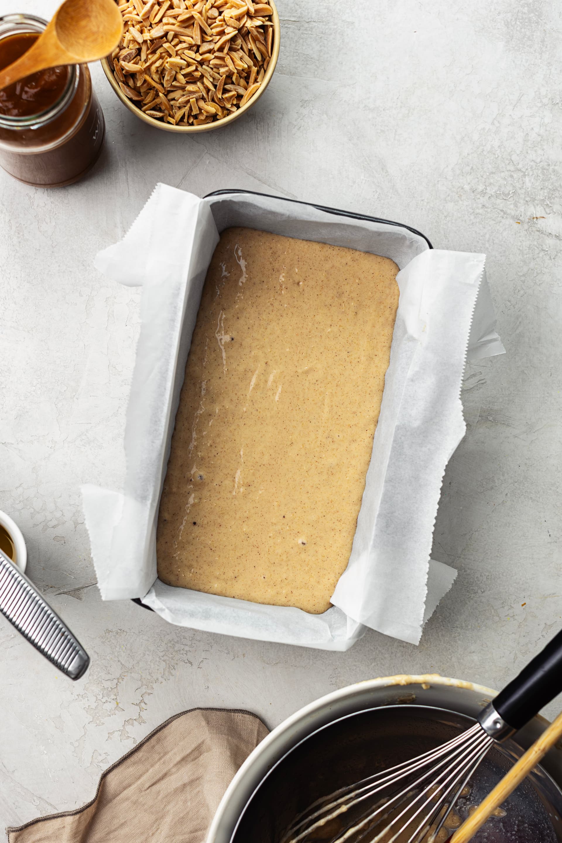 Raw almond cake batter in a parchment-lined tin, with coconut caramel and slivered almonds in the background.