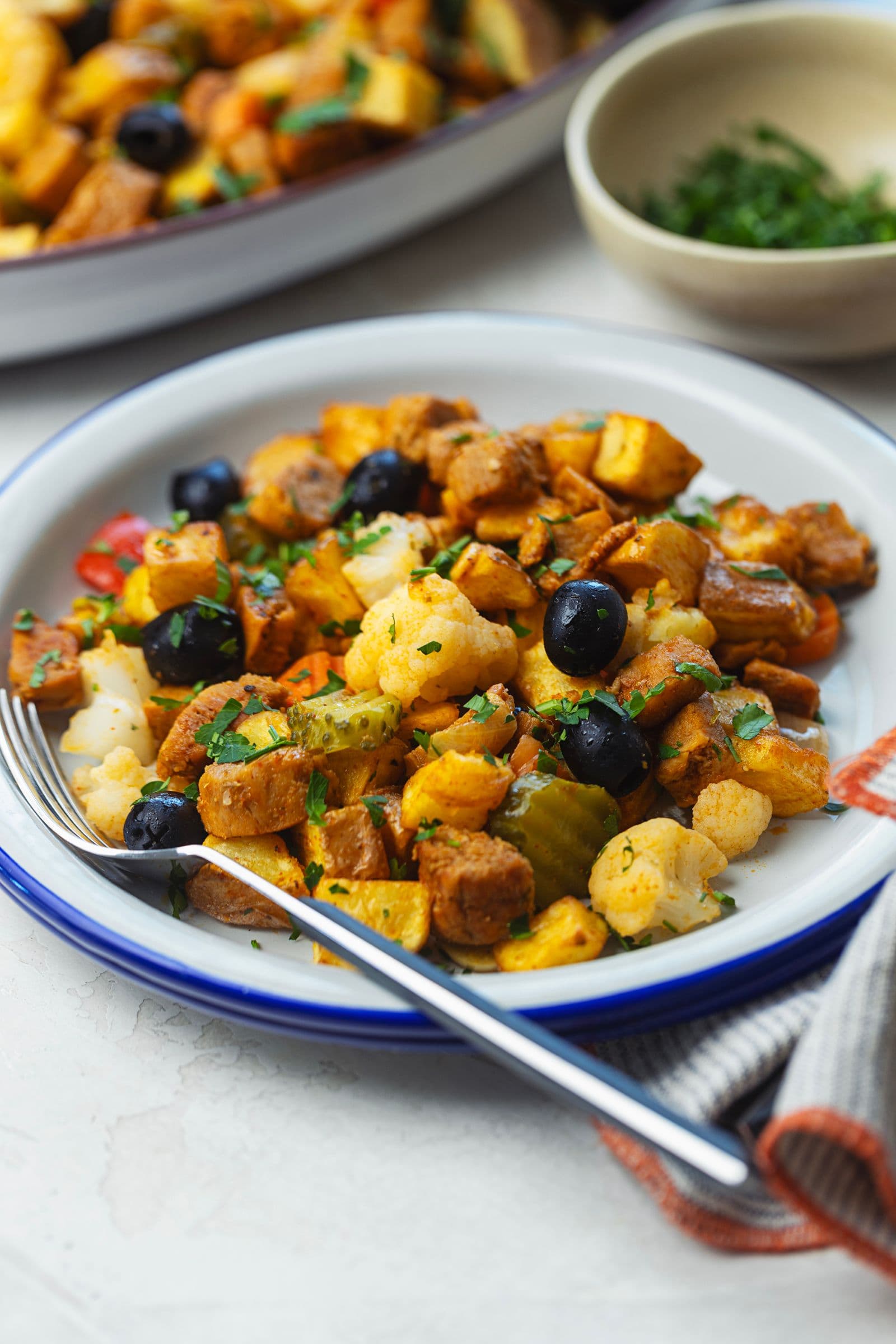Blue-rimmed plate with a portion of soy pica pau at a slightly elevated front angle, showing black olives, pickled cauliflower, crispy potatoes and caramelised soy topped with parsley