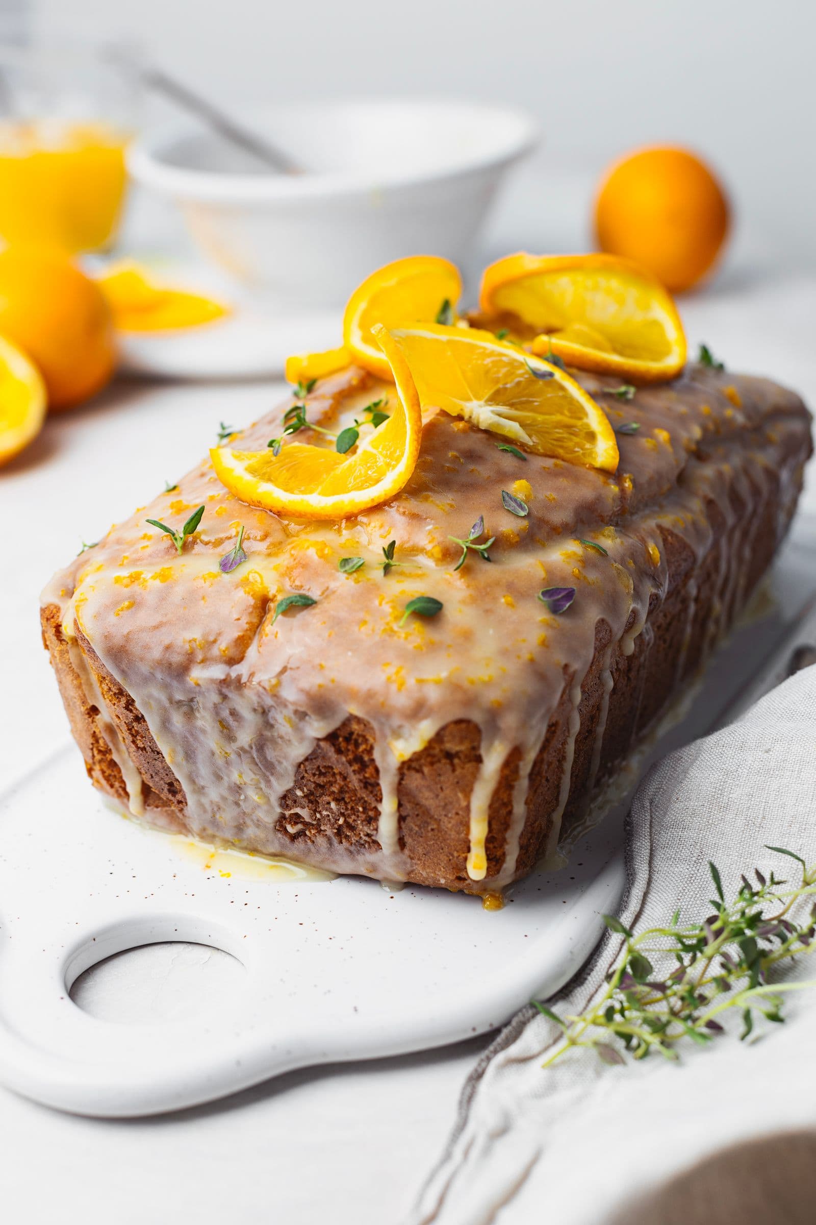 Orange cake with glaze dripping down the sides, topped with orange slices and fresh thyme on a white serving platter.