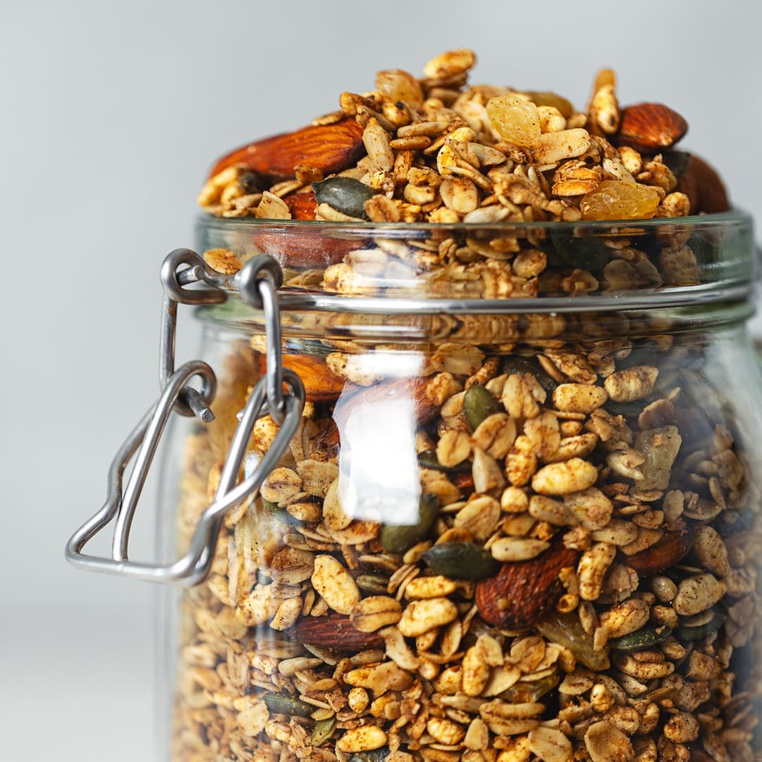 Crunchy chai-spiced granola with almonds, seeds and golden raisins in a glass jar, with some scattered on the table.