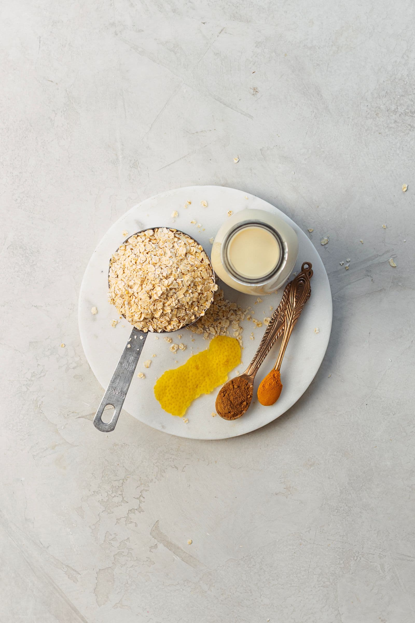 Flat lay of oats, plant milk, lemon peel, cinnamon and turmeric on a white plate over a light grey surface