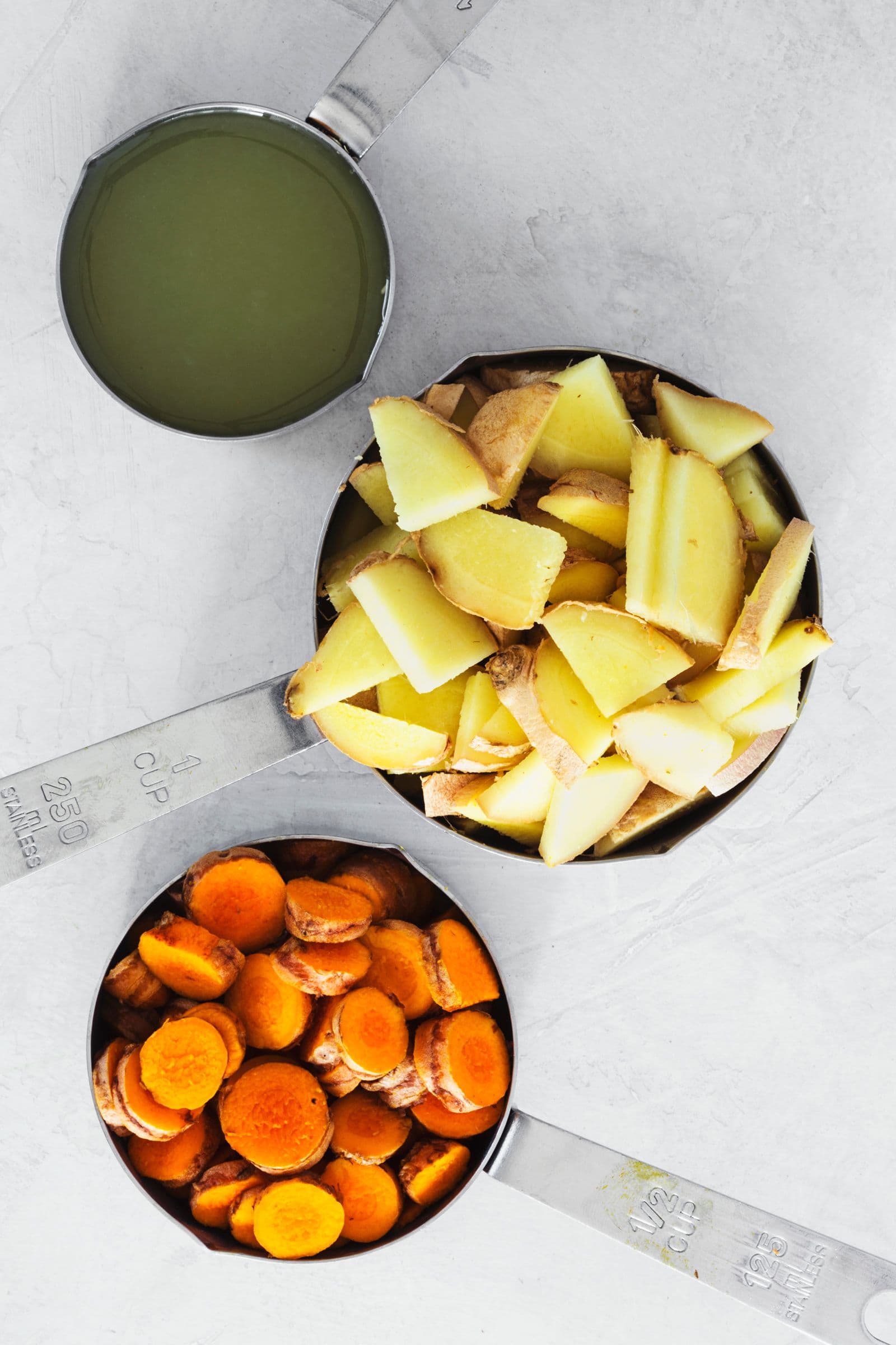 Measuring cups with sliced ginger, turmeric, and fresh lemon juice ready for blending.