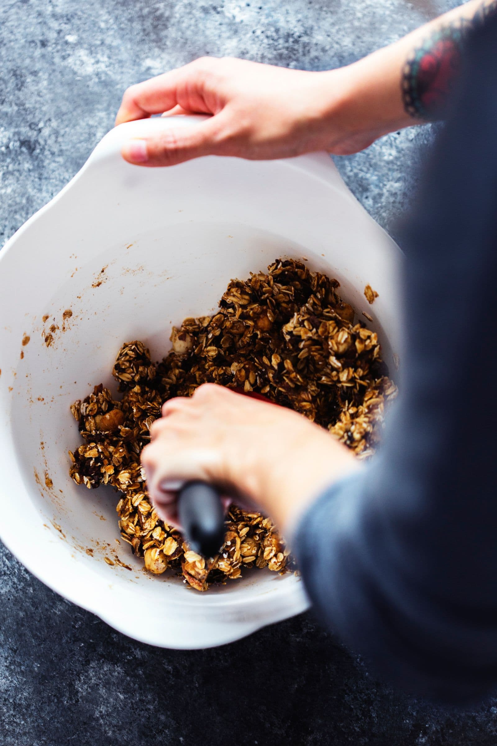 Hands mixing sticky granola dough in a white bowl.