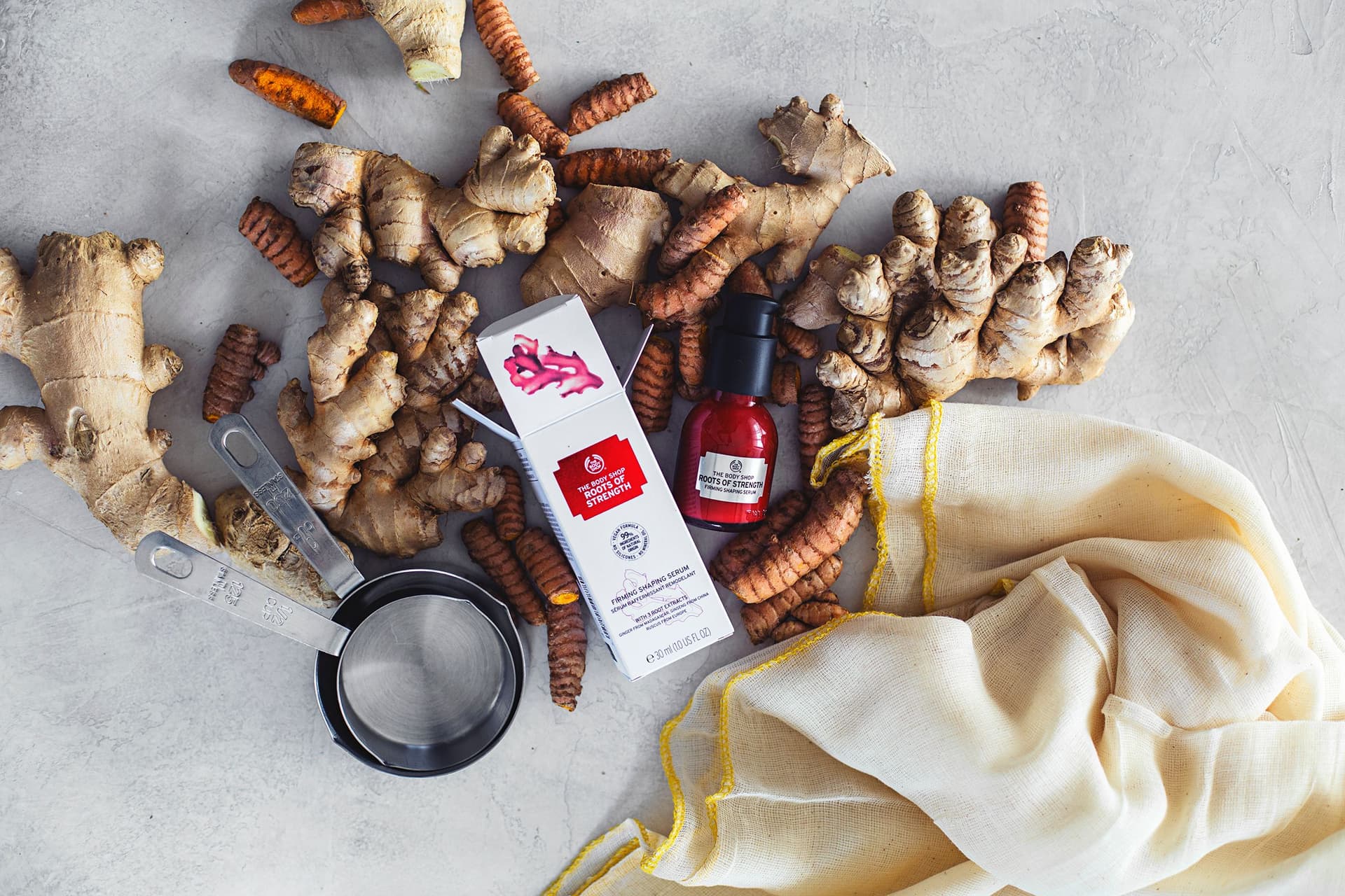 Fresh ginger and turmeric roots with a jar of serum from The Body Shop on a light background.