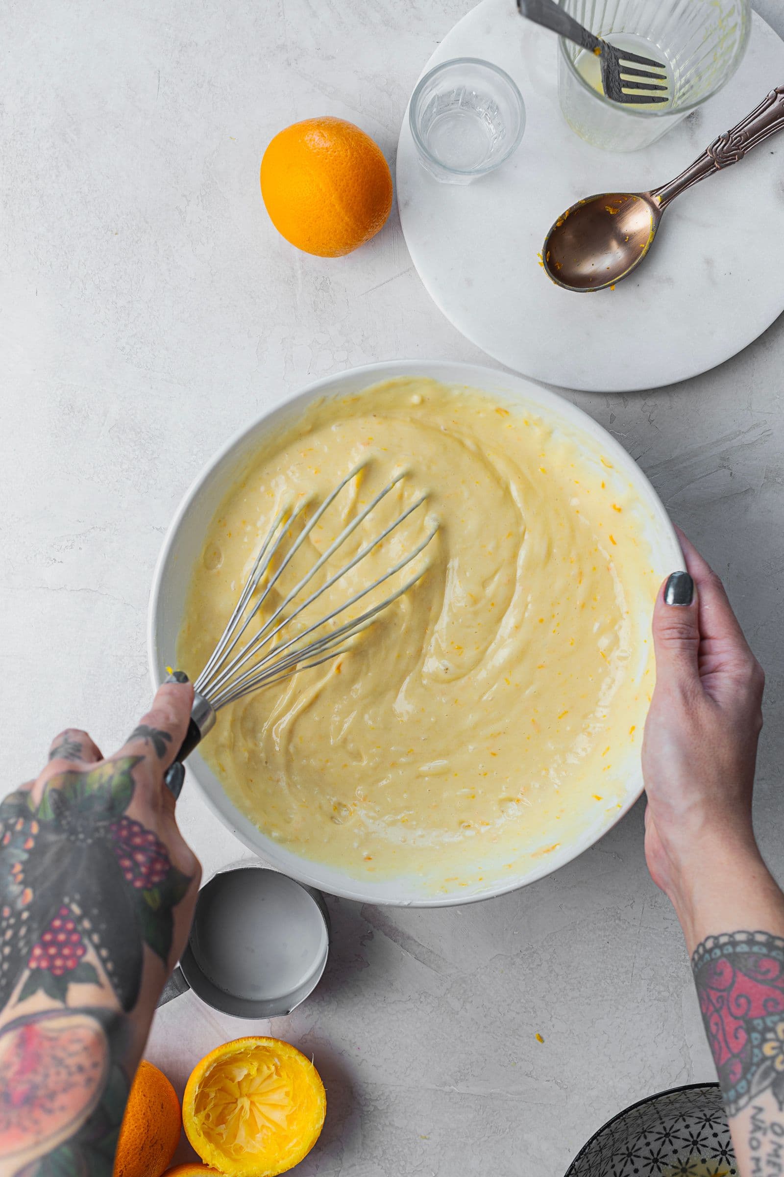 Hands whisking orange cake batter in a white bowl, surrounded by oranges and kitchen utensils.