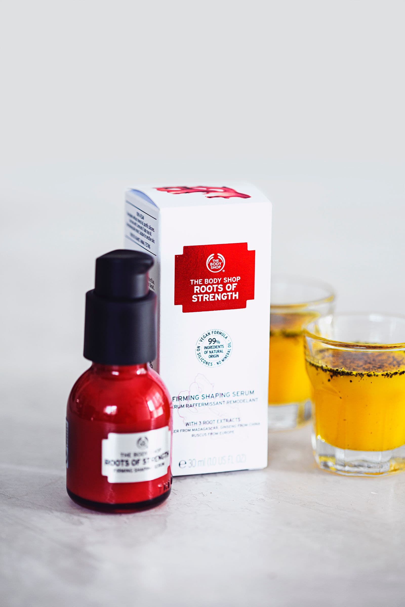 A small bottle of The Body Shop firming serum next to two small glasses of ginger turmeric syrup on white background.