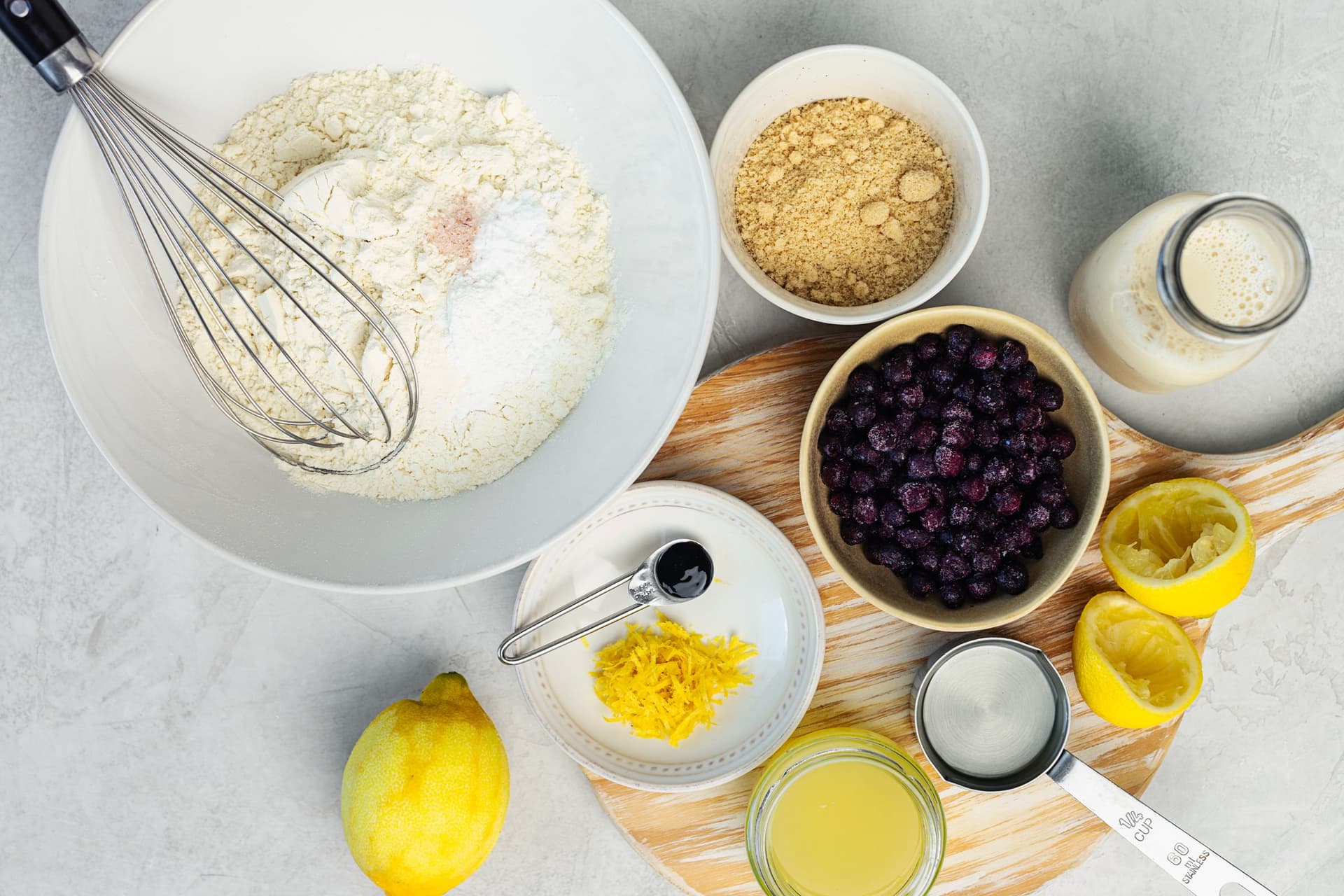 Ingredients for lemon and blueberry streusel muffins laid out on a light surface, including flour, lemon zest, blueberries, plant milk and sugar.