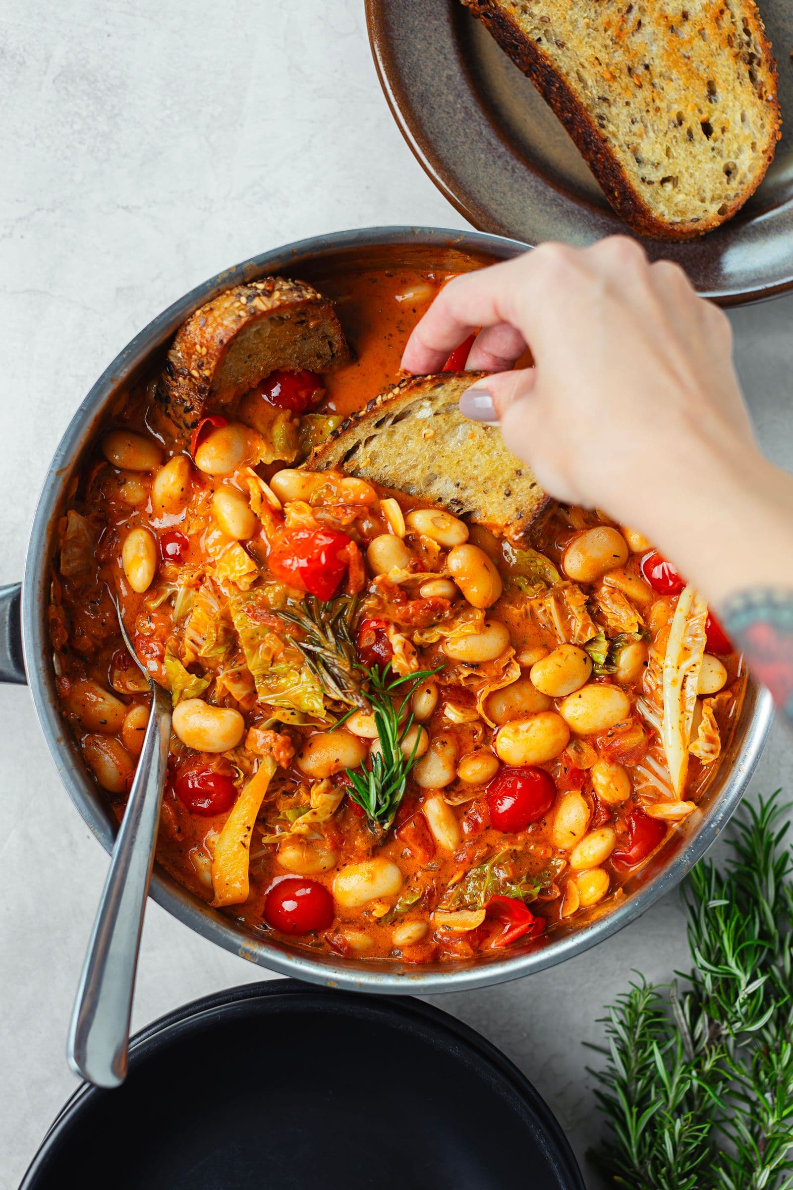 A hand dipping a slice of toasted bread into the pot of hot white bean and cabbage stew.