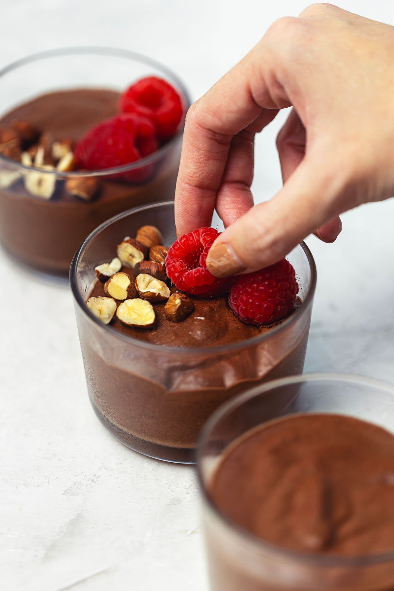 Hand placing fresh raspberries on top of vegan chocolate mousse in an individual glass.